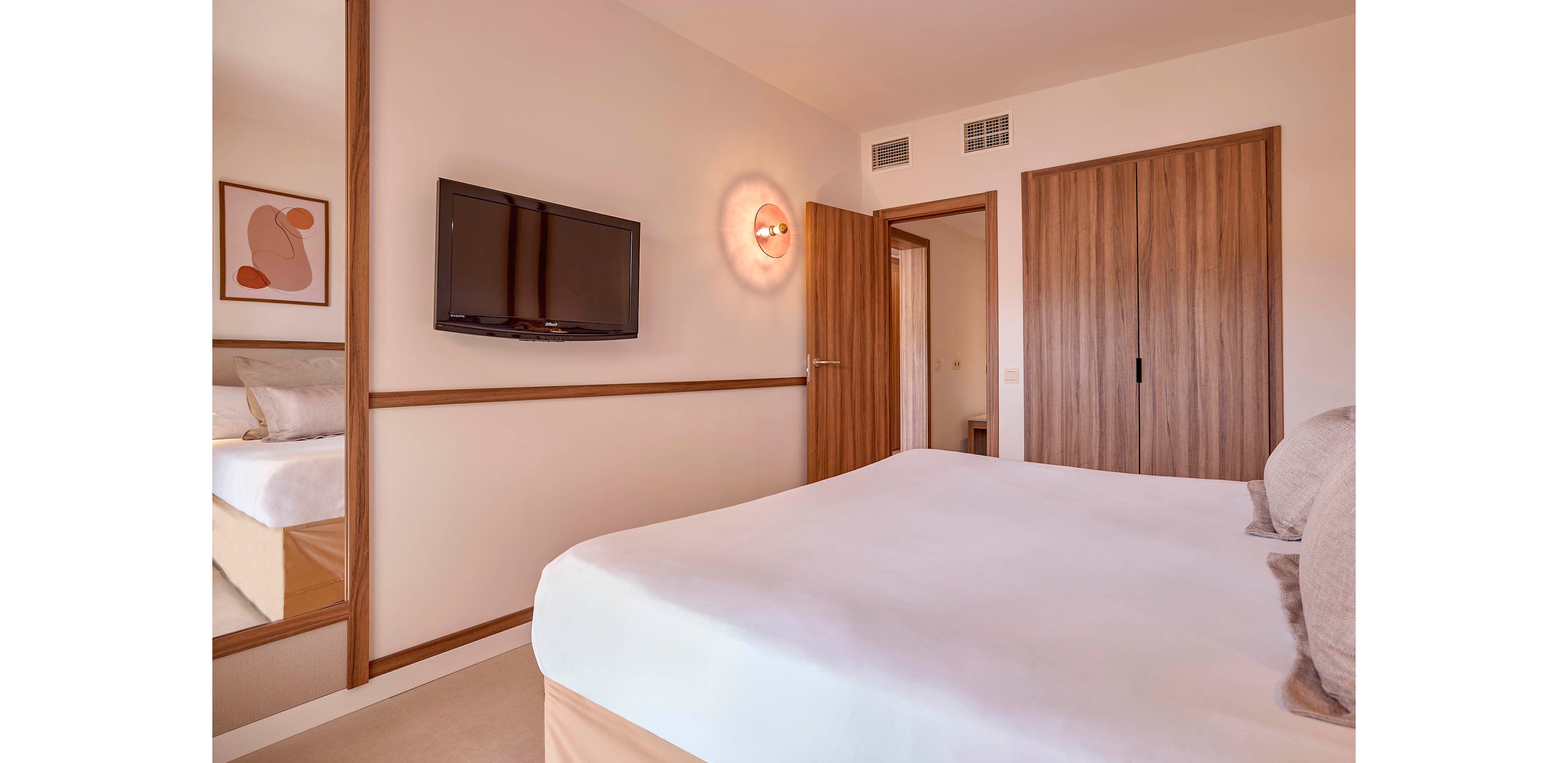 A contemporary hotel bedroom features a neatly made double bed with neutral bedding. A flat-screen television is mounted on the wall opposite the bed, next to a wooden door and built-in wardrobe with wood grain finish. Soft lighting from a wall sconce and natural tones create a calm, minimalist atmosphere. A large mirror and abstract artwork add decorative touches to the clean, modern interior.