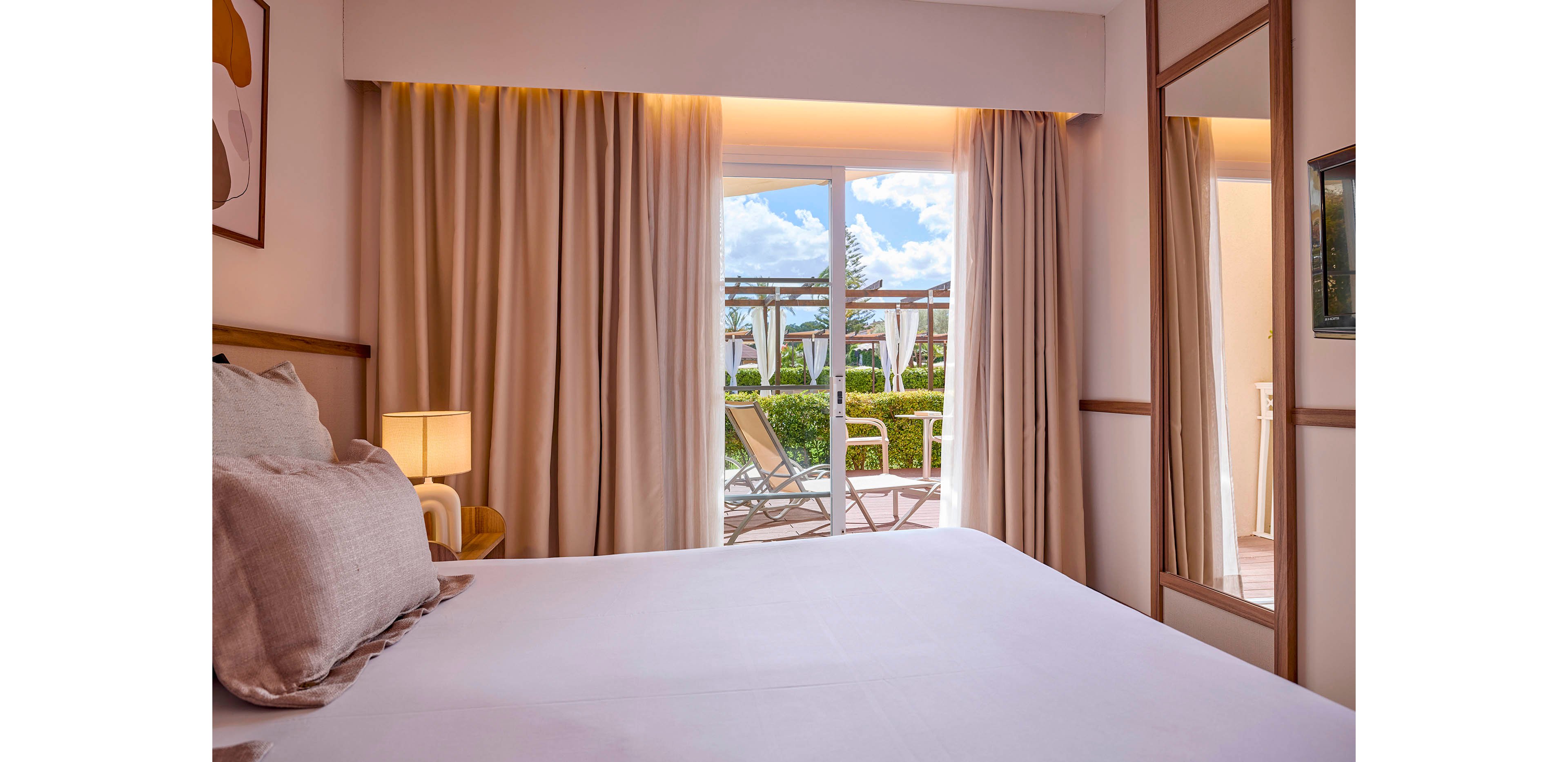 A neatly made double bed sits in a contemporary hotel bedroom with soft neutral tones. Floor-to-ceiling curtains frame a large glass door that opens onto a sunny garden terrace. Outside, lounge chairs and greenery are visible under a blue sky with scattered clouds. Warm ambient lighting and minimal decor create a calm, inviting atmosphere ideal for relaxation.