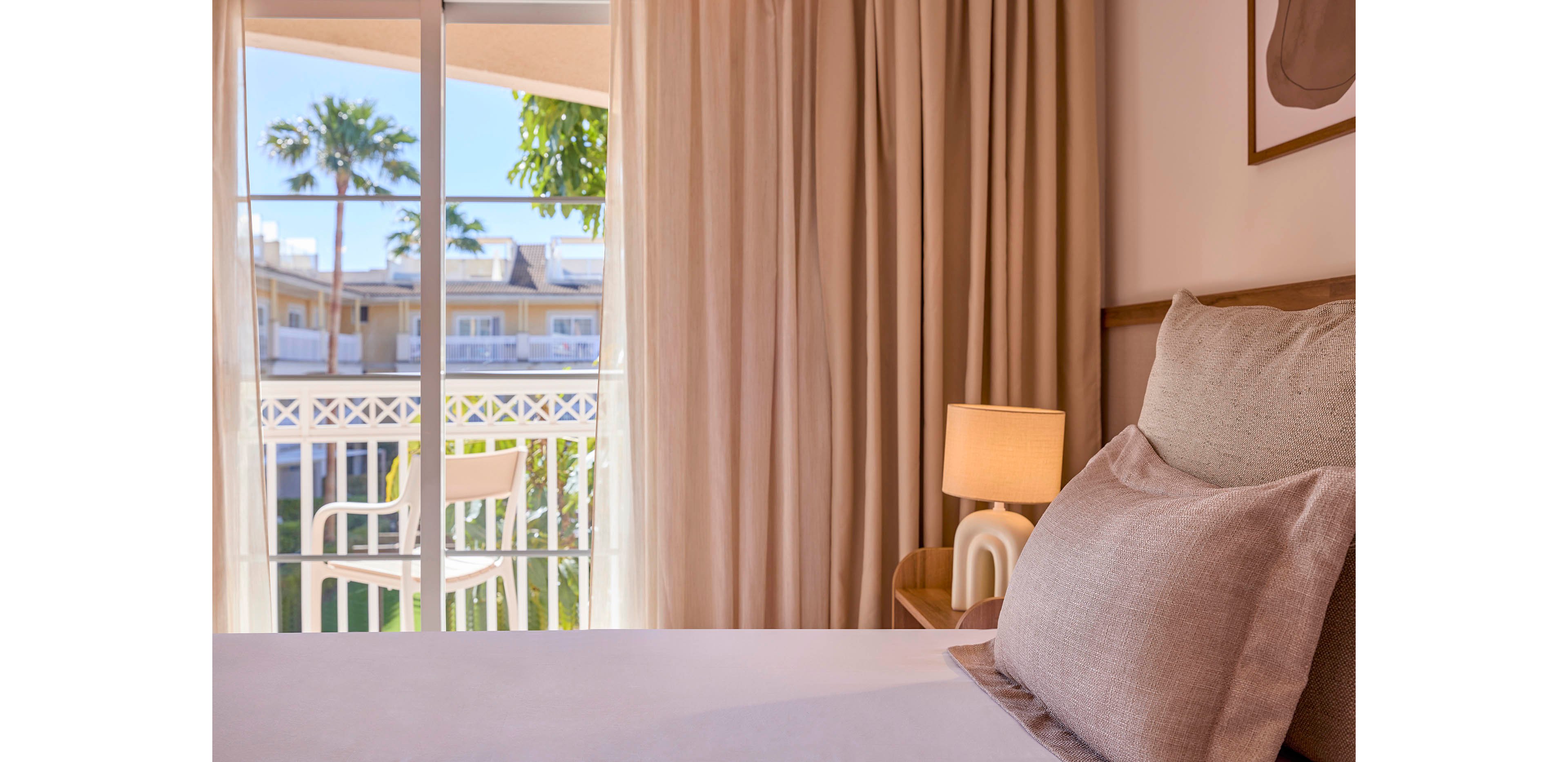 A cozy hotel bedroom features a neatly made bed with soft neutral pillows in the foreground. Floor-to-ceiling curtains frame a glass door that opens onto a small balcony with white chairs and a railing. Warm light from a bedside lamp adds a relaxing ambiance to the room. Outside, palm trees and neighboring buildings suggest a sunny resort or vacation setting.