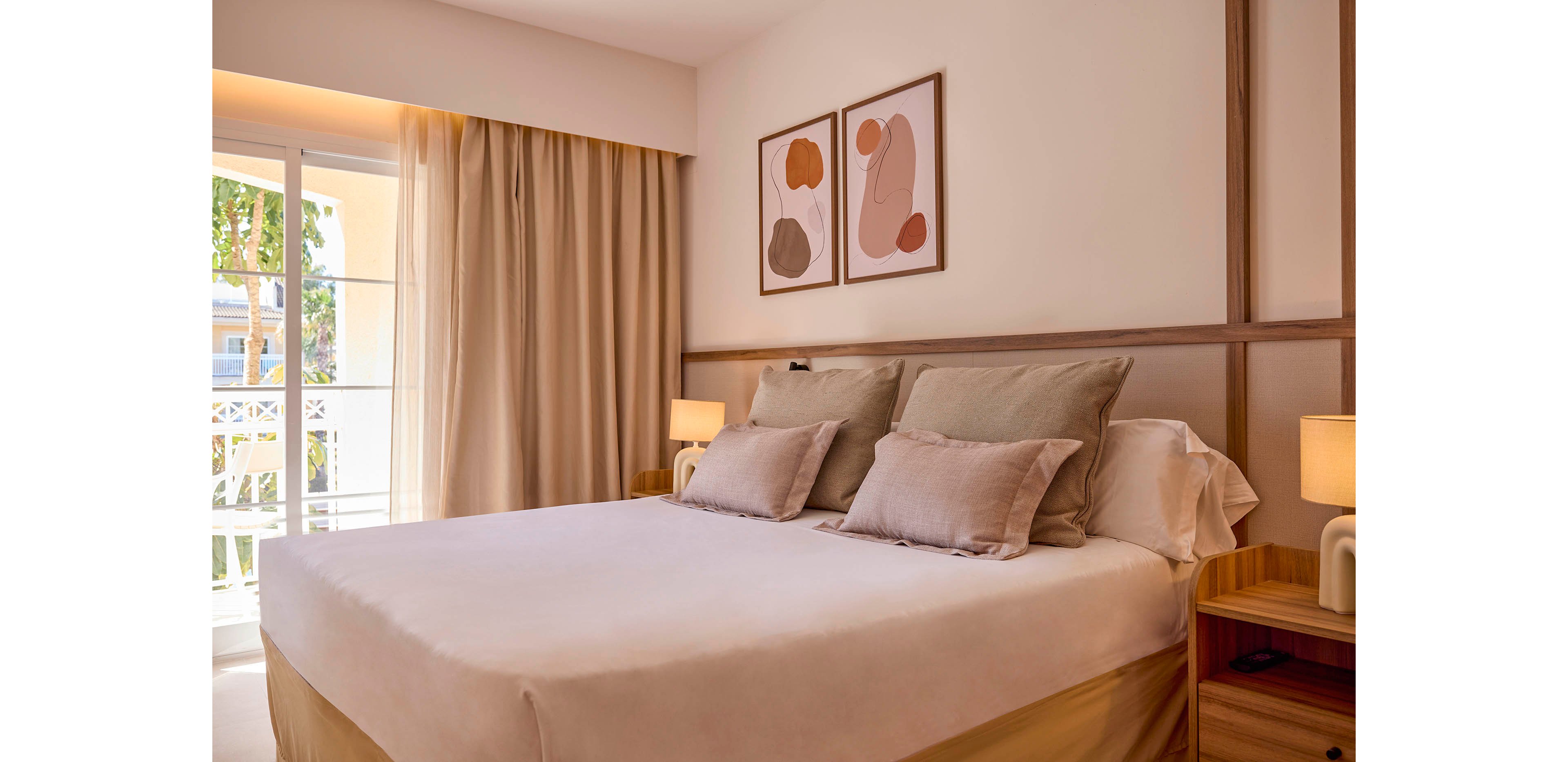 A neatly made double bed with neutral-toned pillows and bedding sits in a modern bedroom. Warm light from bedside lamps and a large window with curtains creates a soft, inviting atmosphere. Minimalist abstract artwork hangs above the headboard, complementing the beige and wood accents. Sunlight filters through the balcony doors, adding a bright, airy feel to the calm interior.