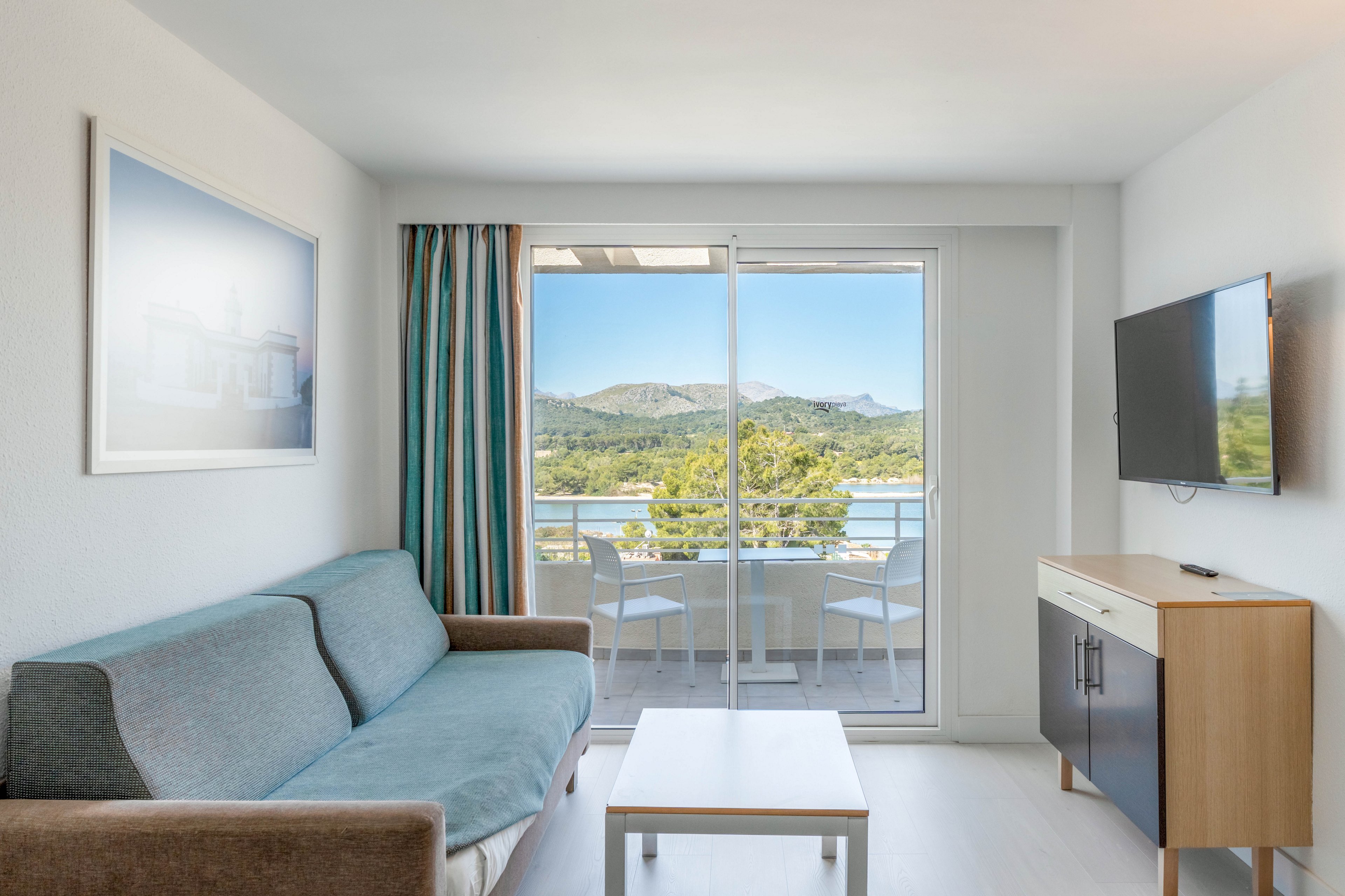 A modern hotel living room features a blue and gray sofa facing a wall-mounted television and a small wooden cabinet. A simple coffee table sits in the center of the light-filled space. Large sliding glass doors open to a balcony with chairs, overlooking a scenic view of trees, water, and distant mountains. Neutral walls, striped curtains, and a framed picture contribute to the clean, minimalist interior design.