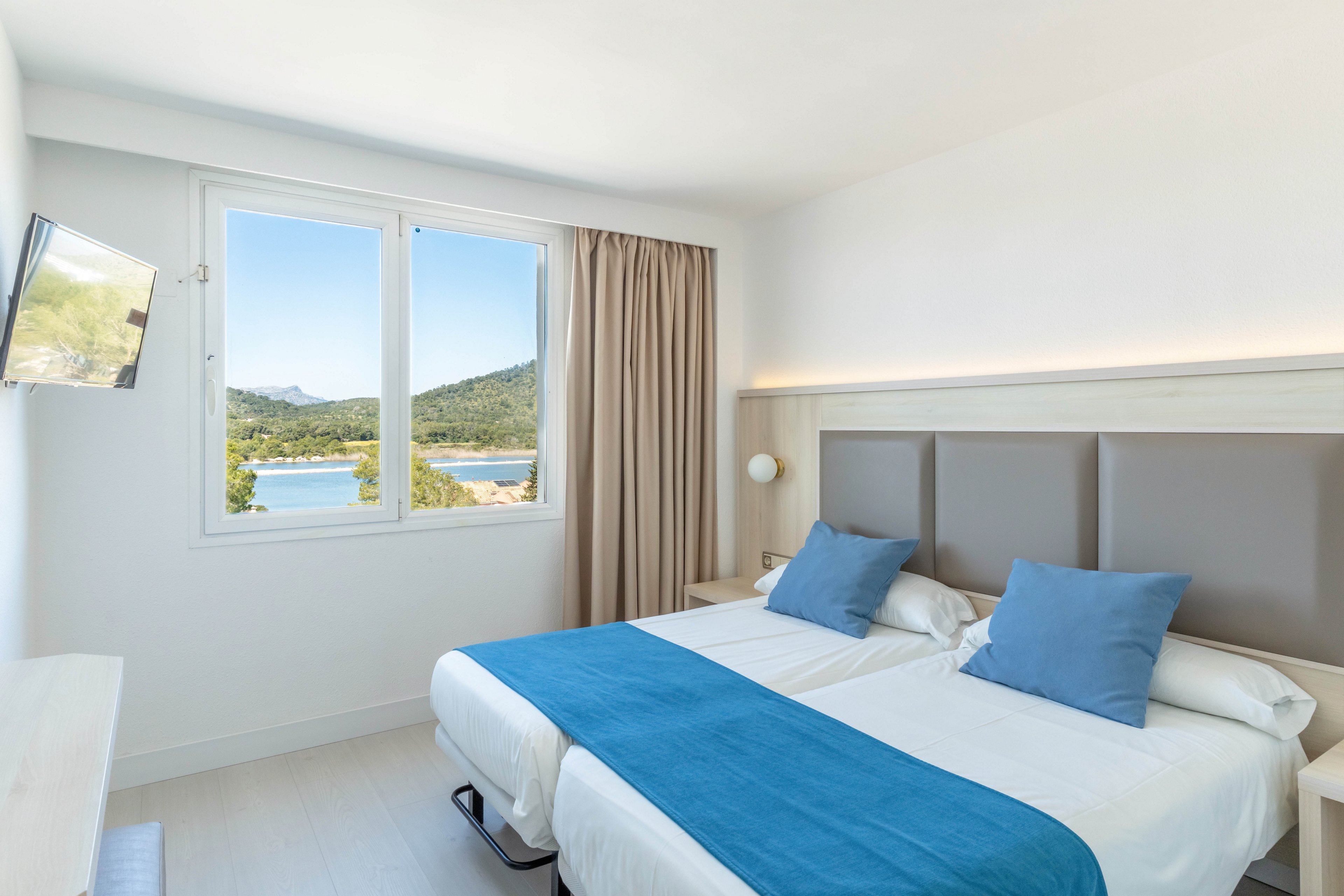 A modern hotel bedroom features two single beds pushed together, dressed in white linens with blue throws and matching pillows. Large windows offer a scenic view of a coastal landscape with water and green hills. Neutral walls, light wood furniture, and a beige curtain create a clean, minimalist aesthetic. A wall-mounted television and bedside lamp complete the contemporary, relaxing space.