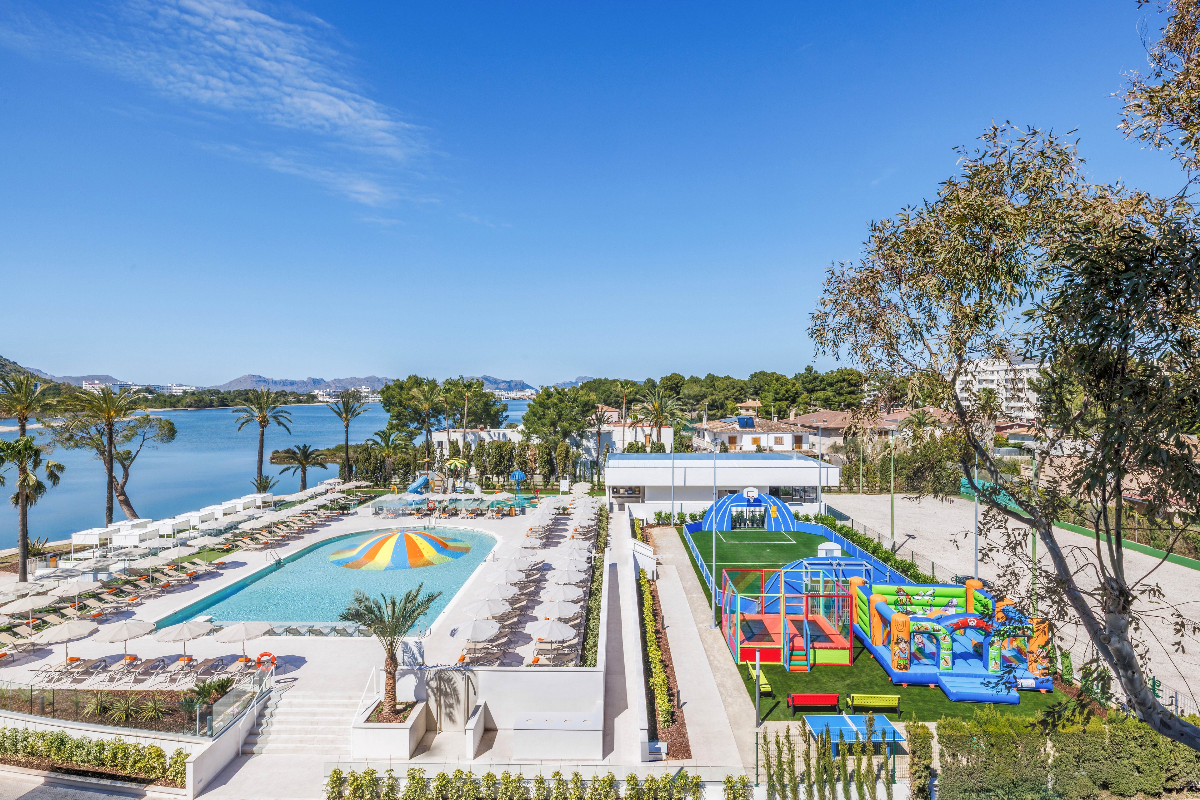 A large outdoor swimming pool with sun loungers overlooks a calm waterfront at a modern resort. Beside the pool, a colorful inflatable playground and play structures sit on bright green artificial grass. Palm trees, landscaped gardens, and low-rise buildings frame the scene under a clear blue sky. The setting suggests a family-friendly vacation destination with dedicated areas for both relaxation and children’s activities.