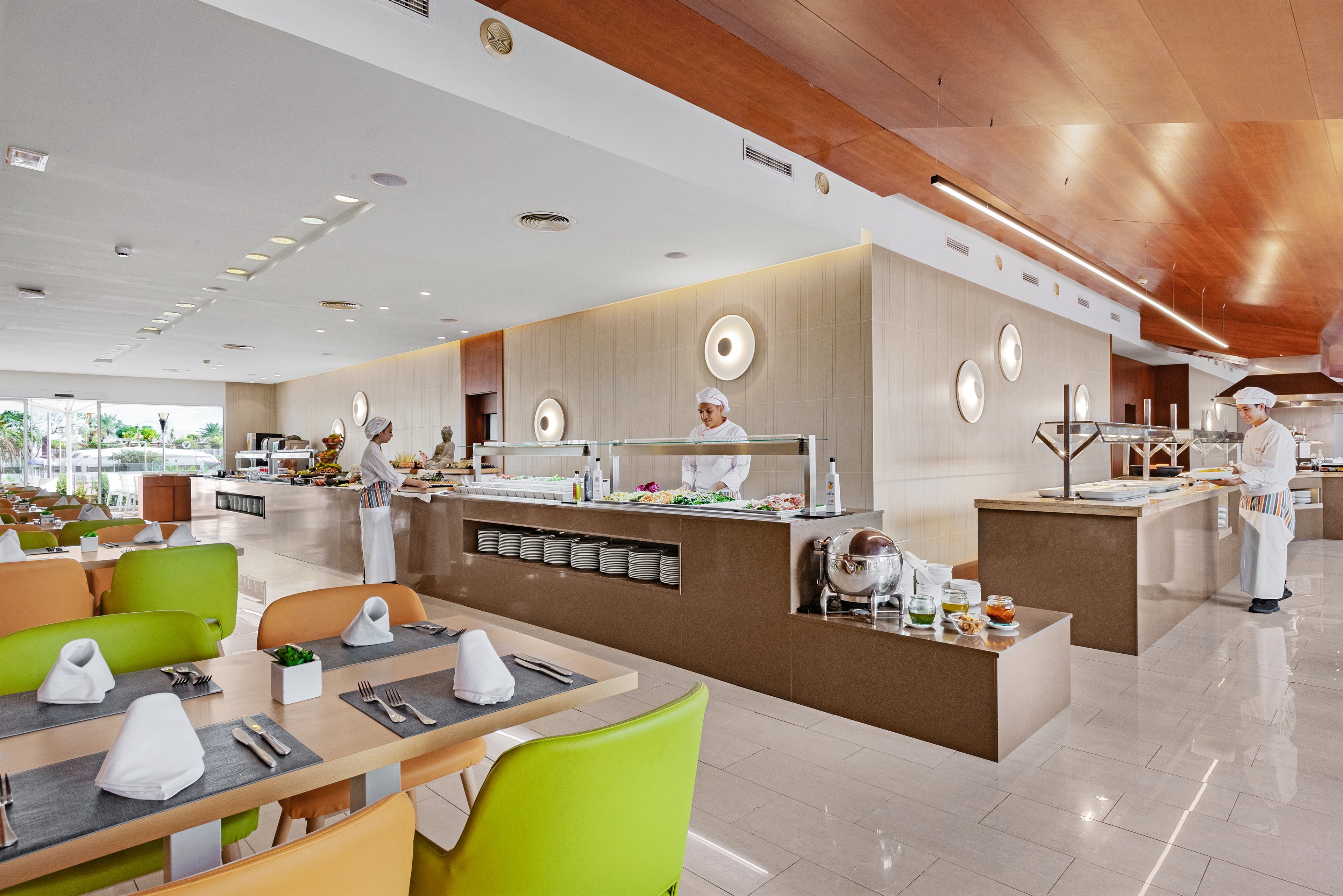 A spacious, contemporary restaurant features a central buffet counter attended by uniformed chefs. Bright green and orange chairs surround neatly set tables with folded napkins and cutlery. The interior design includes warm wood ceilings, light tiled floors, and circular wall lights creating a clean, modern ambiance. Large windows in the background suggest natural daylight and an inviting dining atmosphere.