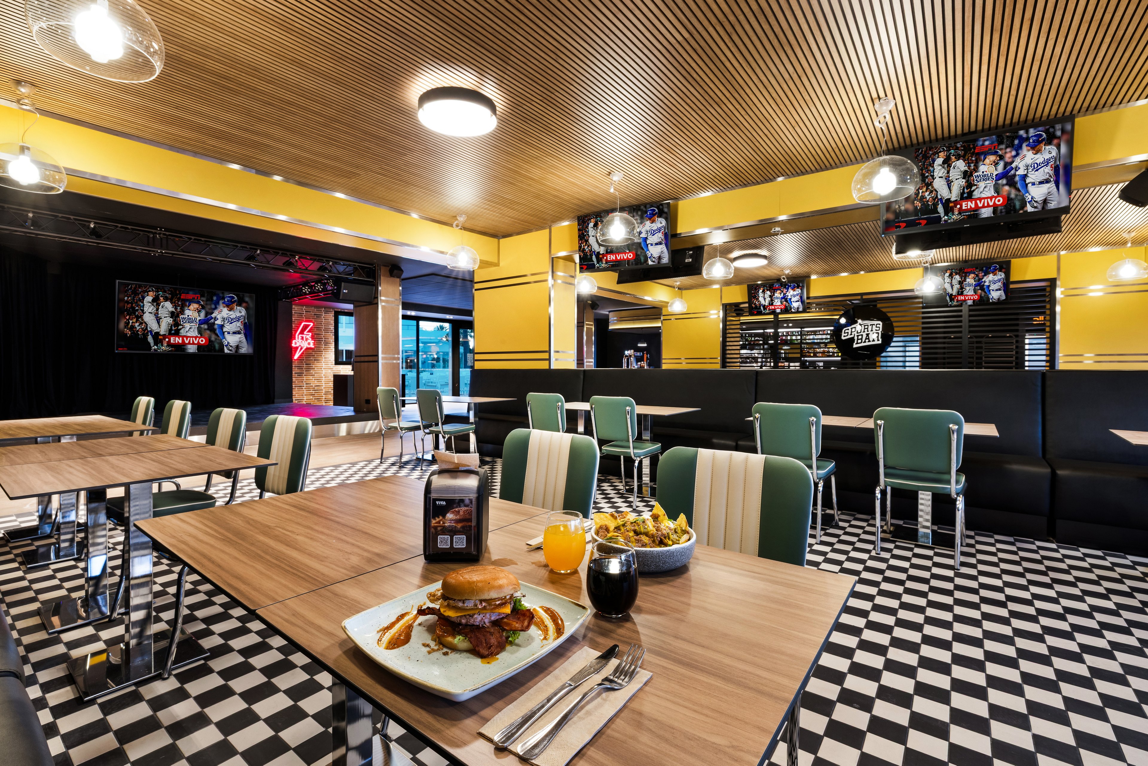 A modern sports bar style diner features checkered black and white flooring and wooden tables with retro green and white chairs. Multiple large flat-screen televisions display sports scenes around the room, creating an energetic viewing atmosphere. In the foreground, a wooden table holds a plated burger with fries and sauce, along with a glass of dark soda and a glass of orange juice. Warm ceiling lights and yellow accent walls contribute to a bright, inviting interior suitable for casual dining and watching games.