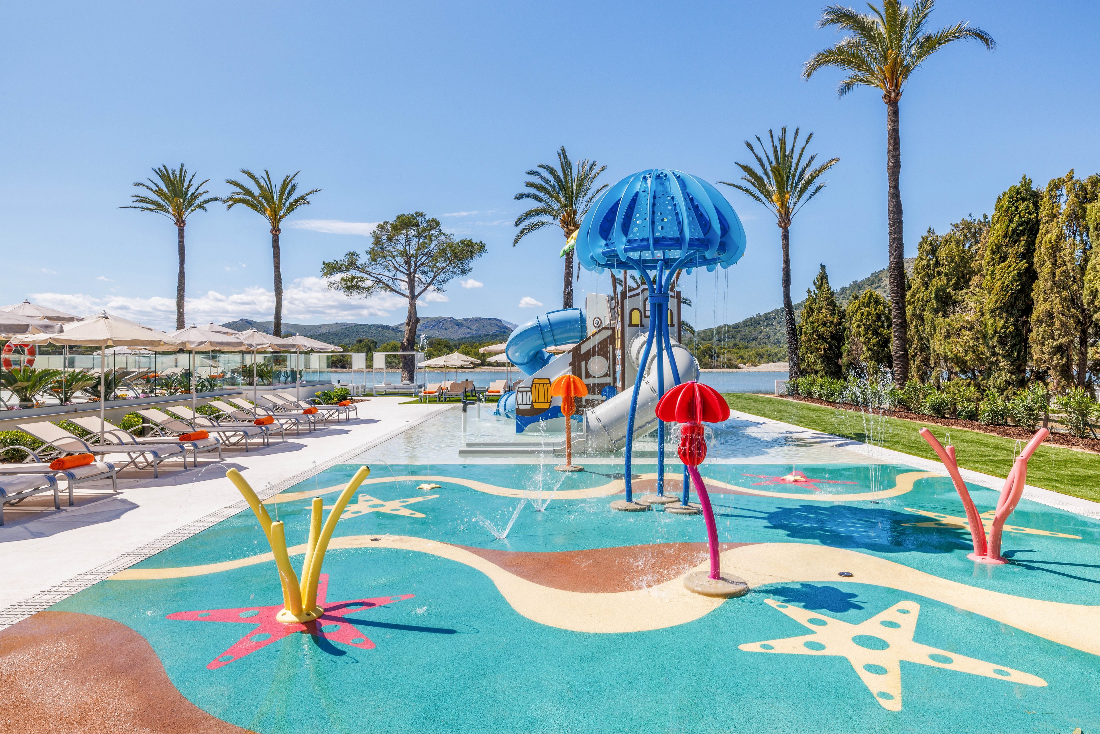 A vibrant children’s splash pad features playful water fountains and slides in a resort pool area. Brightly colored sea-creature structures and starfish patterns decorate the shallow water surface. Sun loungers with umbrellas line the poolside, framed by tall palm trees and manicured greenery. Clear blue skies and distant hills create a sunny, relaxing vacation atmosphere.
