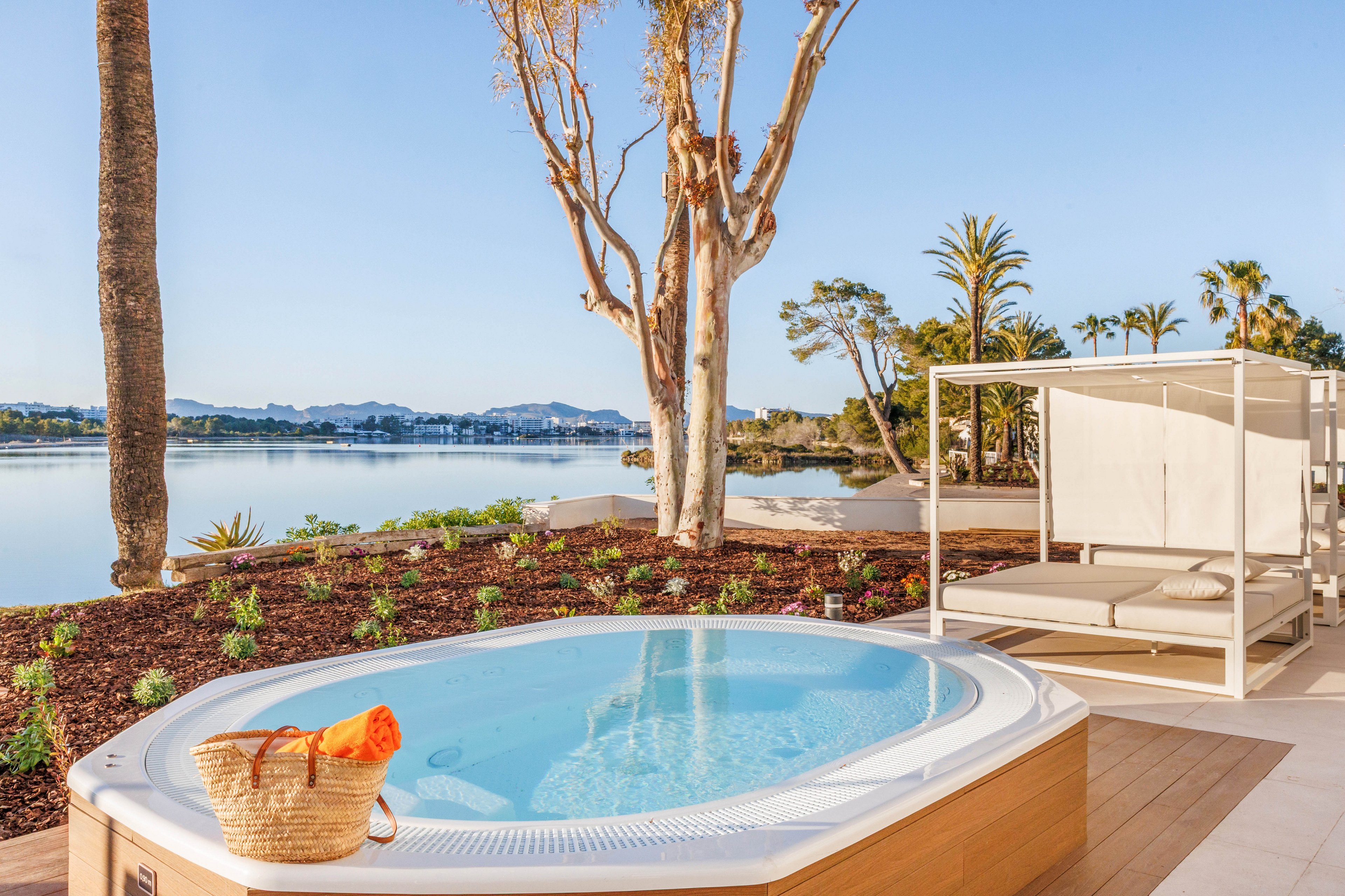 An outdoor hot tub sits on a wooden deck overlooking a calm lake or bay. A modern white canopy daybed is positioned beside the tub, offering a shaded lounging area. Palm trees and landscaped plants surround the space, with distant hills and waterfront buildings visible across the water. A woven beach bag with an orange towel rests on the tub’s edge, enhancing the relaxed vacation atmosphere.