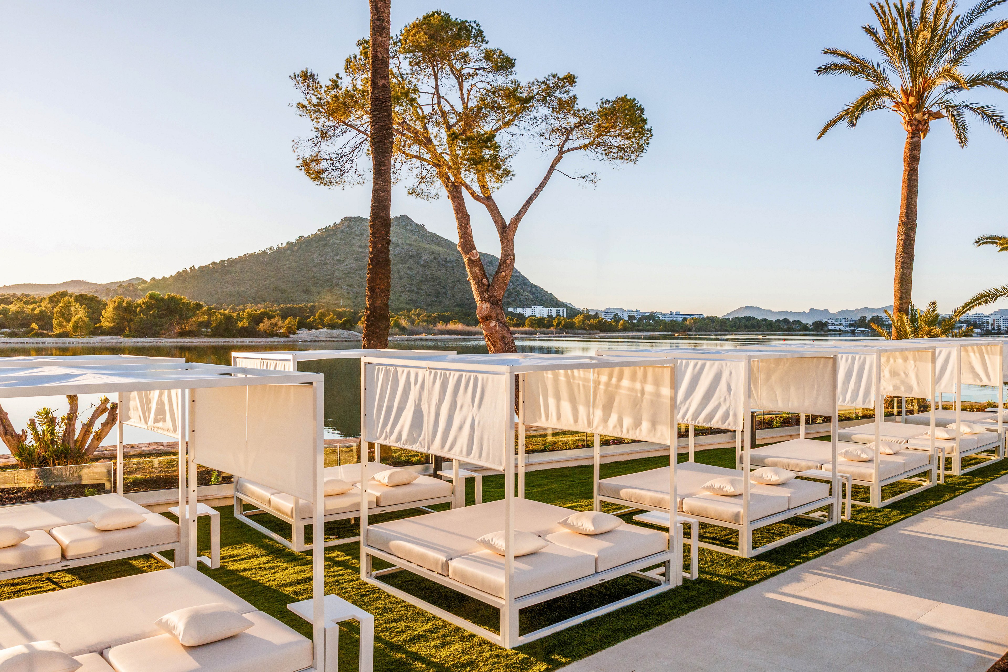 A row of modern white cabanas with cushioned loungers lines a manicured lawn beside a calm lake. The scene is set at sunset, casting warm golden light over the water and surrounding landscape. A tree and tall palm trees frame the view, with distant hills and low buildings visible across the lake. The overall atmosphere is tranquil and luxurious, suggesting a high-end resort or spa retreat.