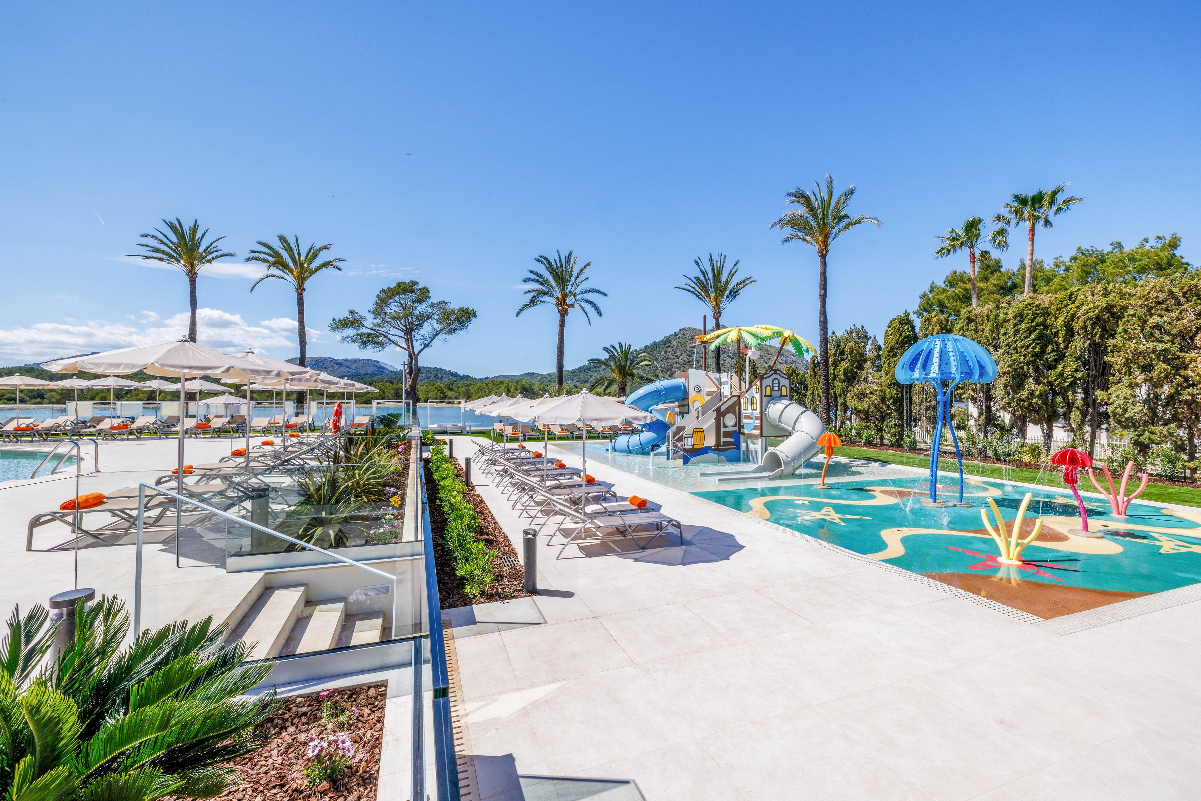 An outdoor resort pool area is shown under a clear blue sky, featuring lounge chairs and umbrellas neatly arranged along the water. Beside the main swimming pool, a colorful children's water playground includes slides, fountains, and mushroom-shaped sprayers. Tall palm trees and lush greenery frame the scene, enhancing the tropical vacation atmosphere. The bright sunlight and vivid colors create a cheerful, family-friendly setting ideal for relaxation and play.