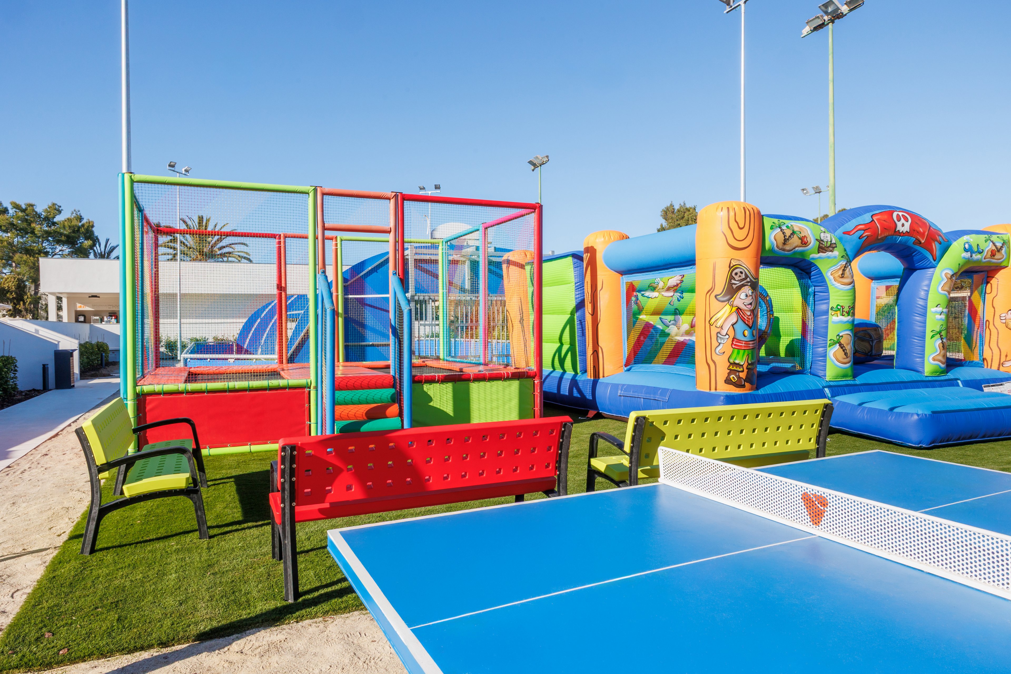 An outdoor recreation area features a bright blue table tennis table in the foreground. Behind it, colorful inflatable bouncers and a trampoline-style play structure create a lively playground scene. Red and green benches line the artificial grass, offering seating for visitors. The setting is sunny and open-air, with tall light poles suggesting a sports or leisure complex.
