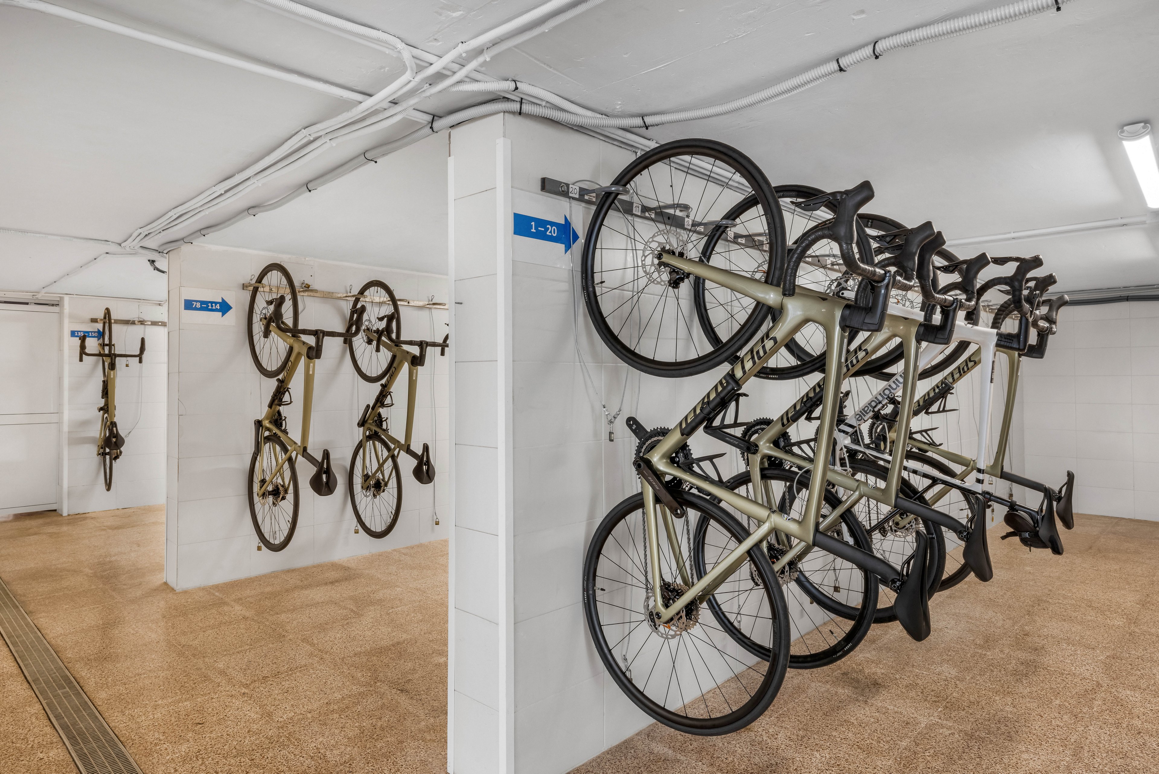 Several bicycles are stored vertically on wall-mounted racks in a clean indoor room. White tiled walls and a light brown floor create a bright, organized environment. Blue directional signs with white arrows are mounted on the walls to guide users. The space appears to be a dedicated bike parking area, likely in a residential or commercial building.