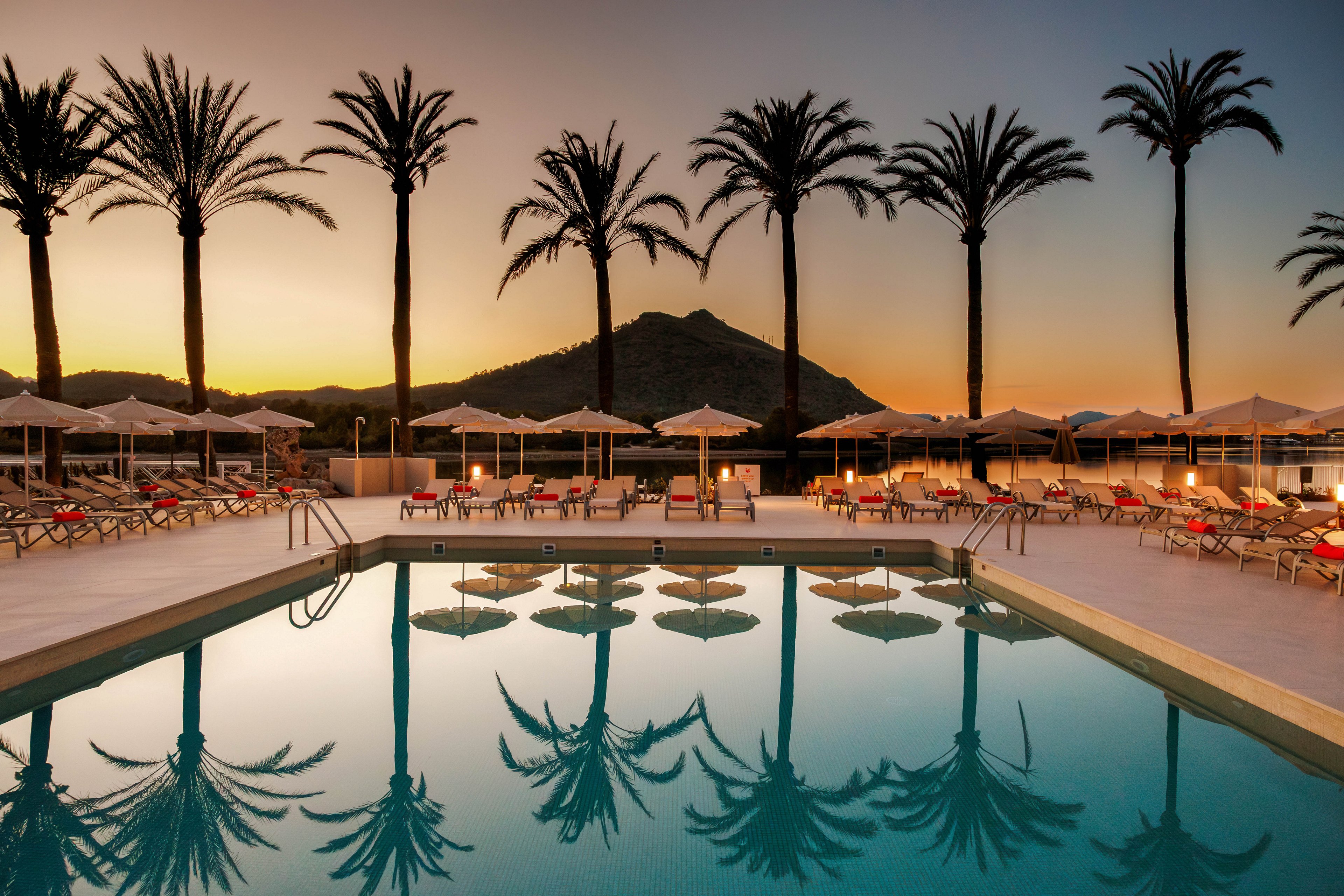 An inviting resort swimming pool reflects tall palm trees at sunset. Neatly arranged sun loungers and umbrellas line the poolside, suggesting a tranquil vacation setting. A mountain silhouette rises in the background beneath a warm, colorful sky. The calm water and empty seating create a peaceful, relaxing atmosphere.