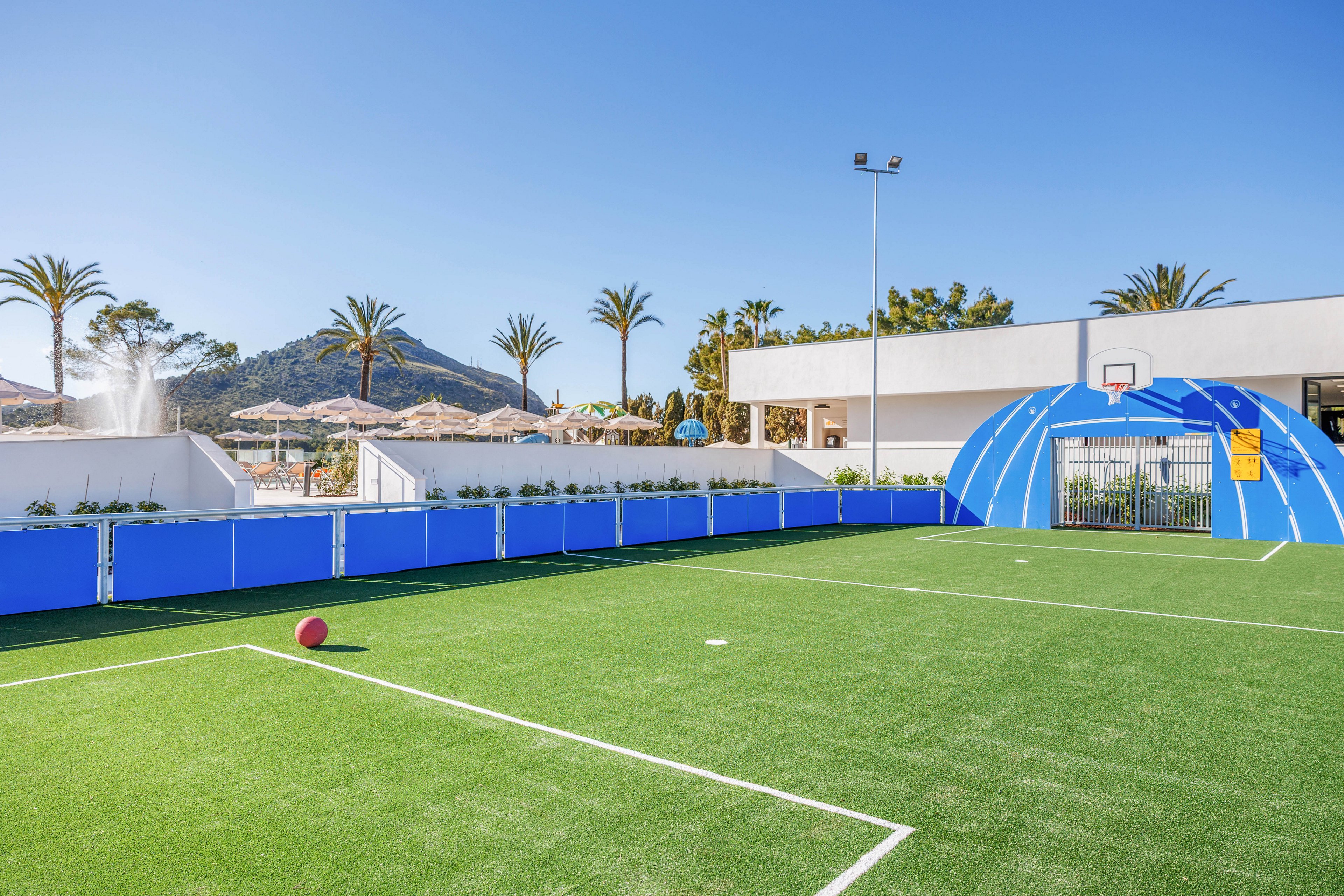 An outdoor sports court with bright green artificial turf sits under a clear blue sky. A red ball rests on the field near white boundary lines, and a small basketball hoop is mounted above a blue tunnel-like structure. Modern white buildings and blue barriers surround the court, with palm trees and a mountain visible in the background. A water fountain and sun umbrellas suggest a resort or recreational setting.