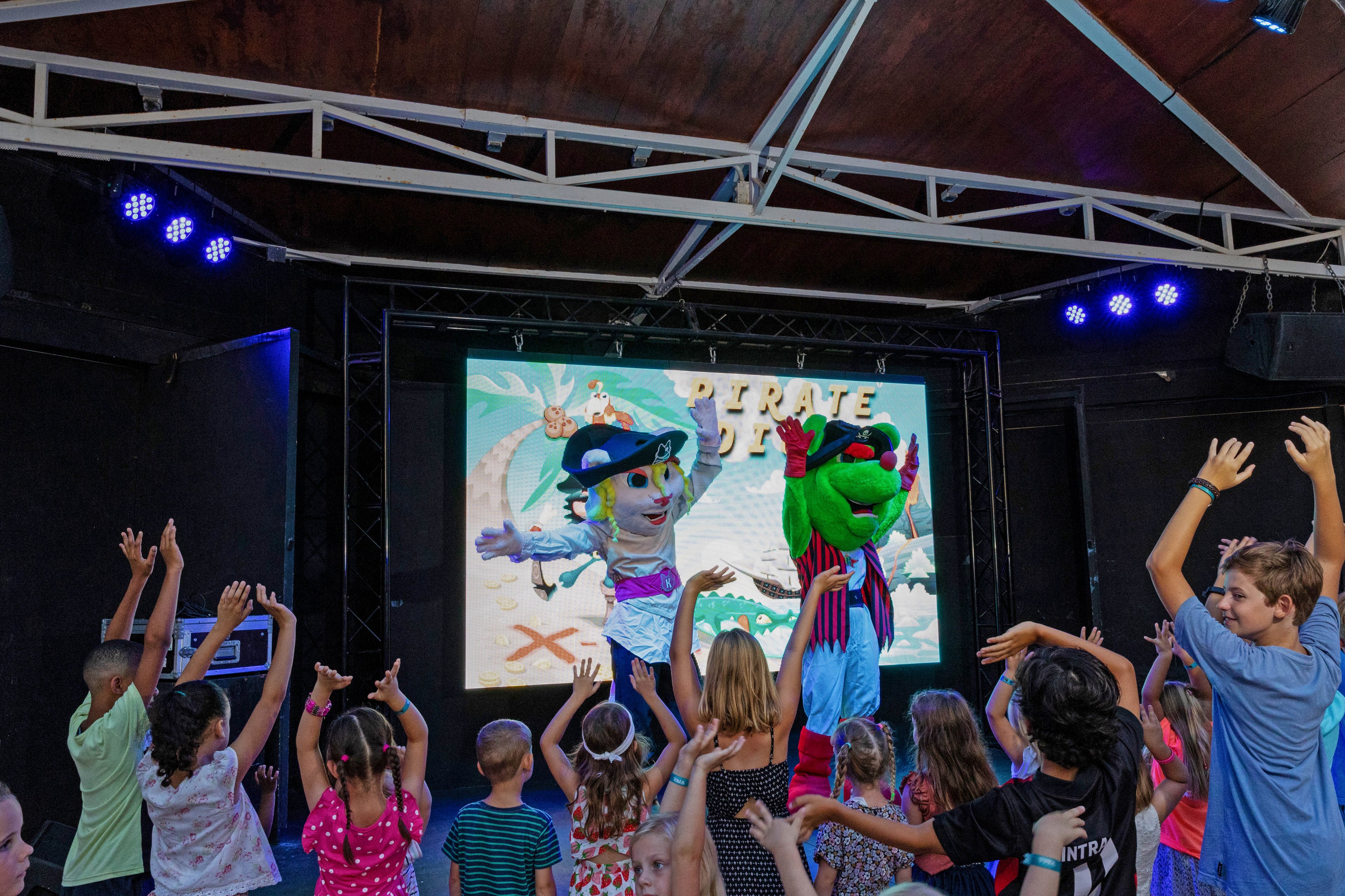 A group of children stand in front of a stage, dancing with their arms raised toward two large costumed mascots. The performance takes place indoors under stage lighting, with a bright digital screen in the background showing a pirate-themed graphic. The kids appear to be enjoying an interactive show or party activity. Vivid colors, playful characters, and energetic poses create a festive, family-friendly atmosphere.