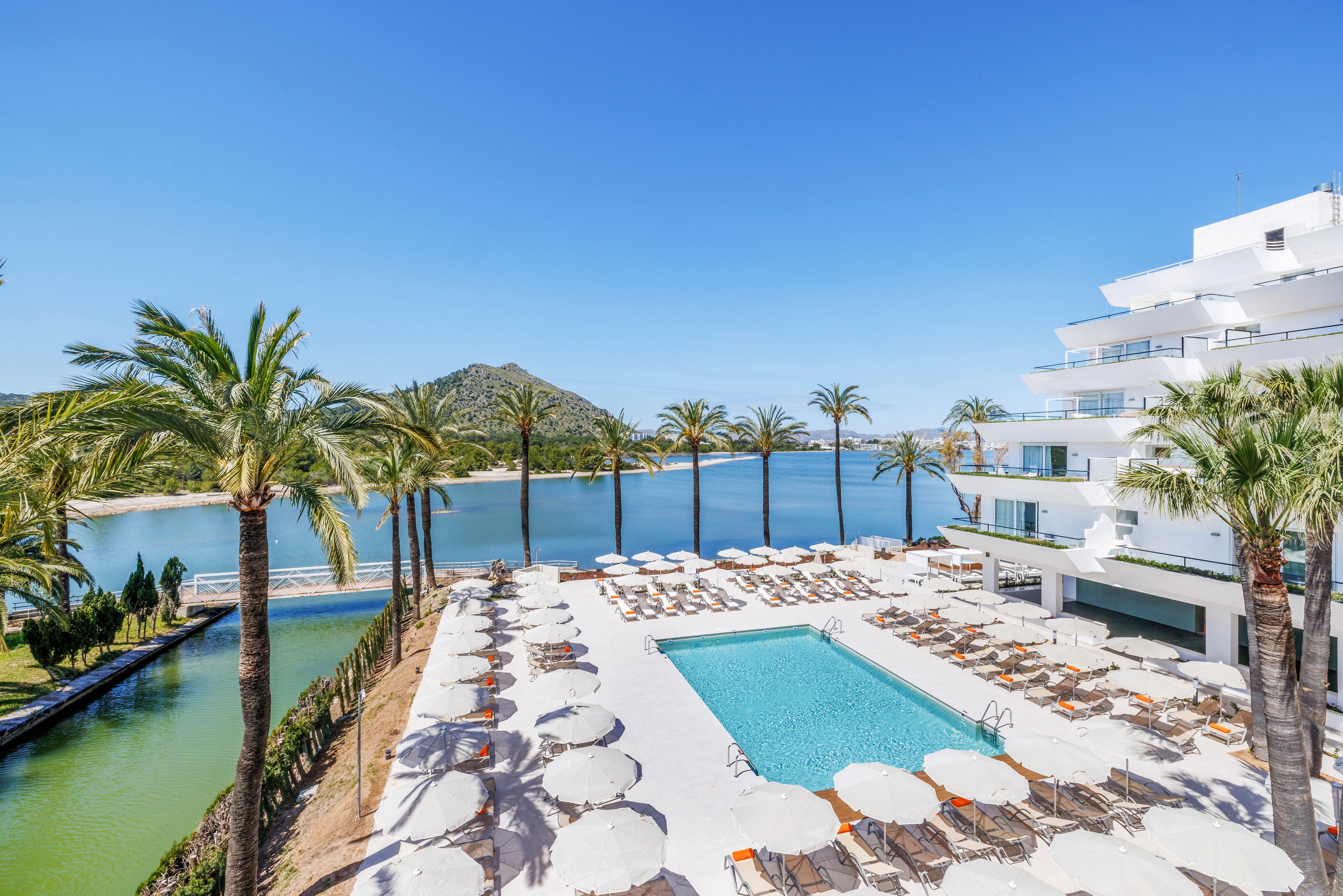 A modern seaside resort features a large rectangular outdoor swimming pool surrounded by sun loungers and umbrellas. Tall palm trees line the terrace, creating a tropical atmosphere next to a calm body of water. In the background, a mountainous coastline rises under a clear blue sky. The white multi-story hotel building overlooks the pool area and the tranquil bay.