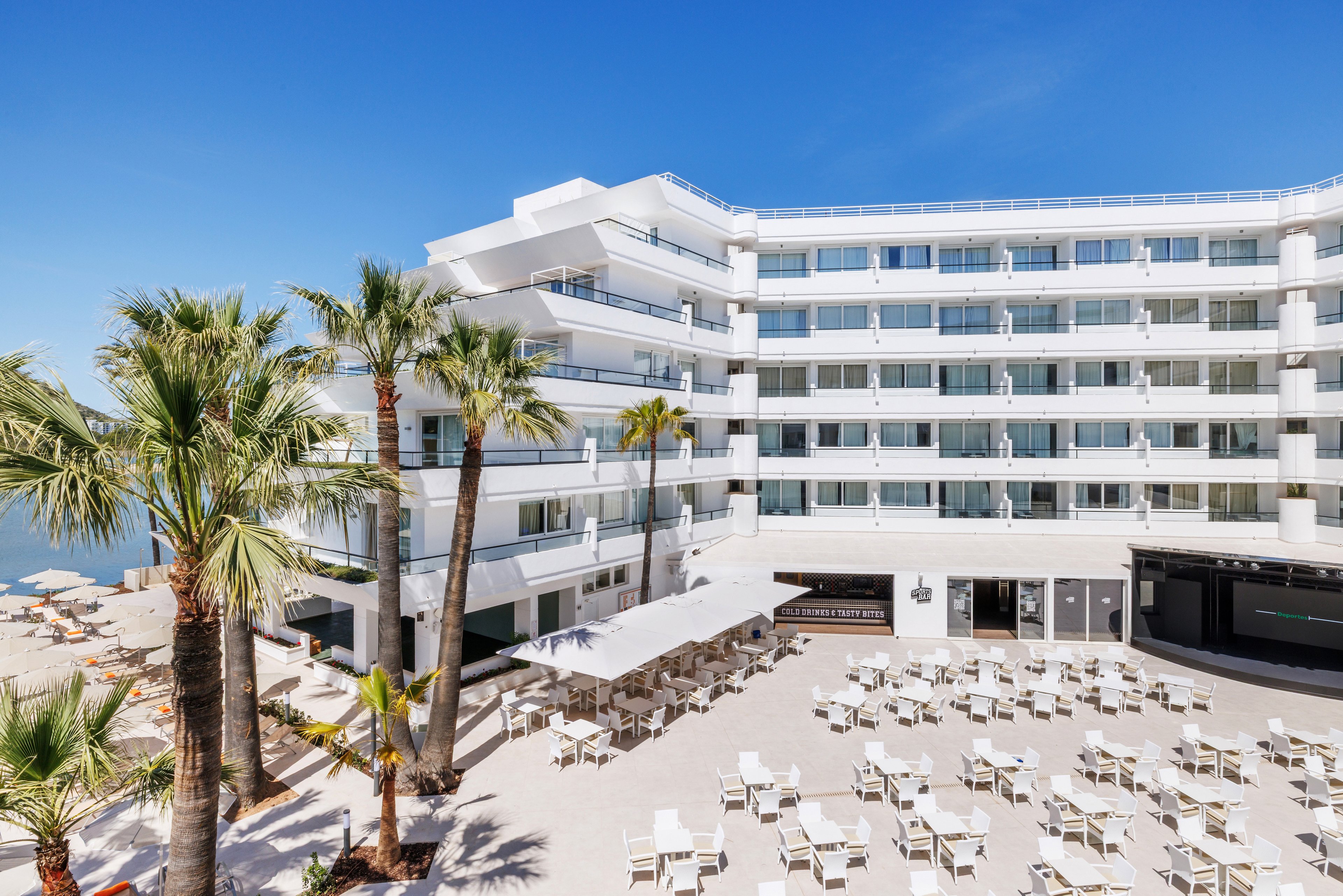 A contemporary multi-story hotel stands beside a sandy beachfront under a clear blue sky. Numerous white tables and chairs are arranged on a spacious outdoor terrace, suggesting a dining or event area. Tall palm trees frame the scene, adding a tropical atmosphere. The building’s clean white facade and glass balconies create a bright, modern resort feel.