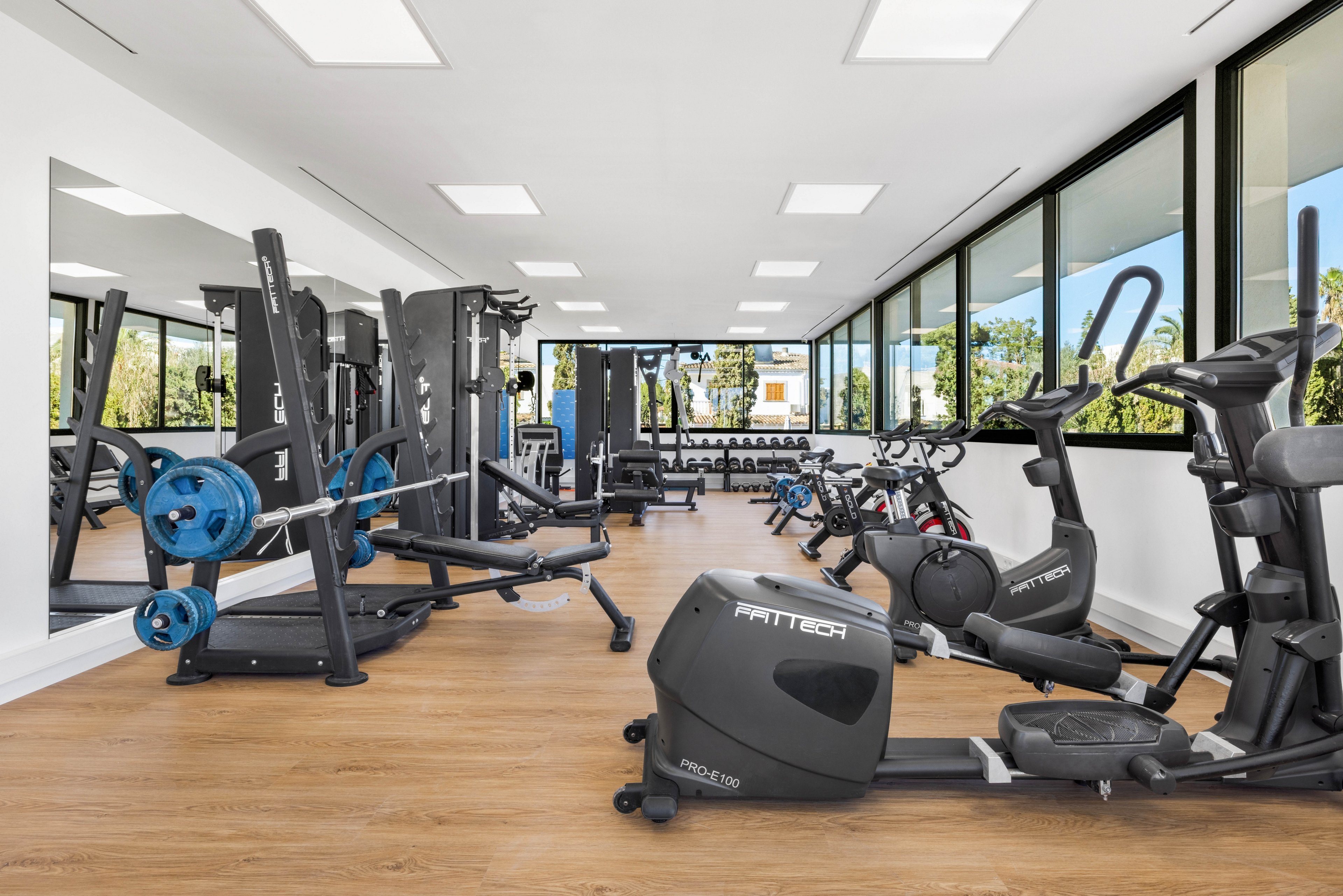 A spacious, modern gym interior features various strength and cardio machines neatly arranged on a light wood floor. Large windows along one wall let in abundant natural light and offer views of greenery outside. Stationary bikes, weight benches, and resistance machines are visible, with mirrors enhancing the sense of space. The room appears clean, organized, and ready for individual workouts.