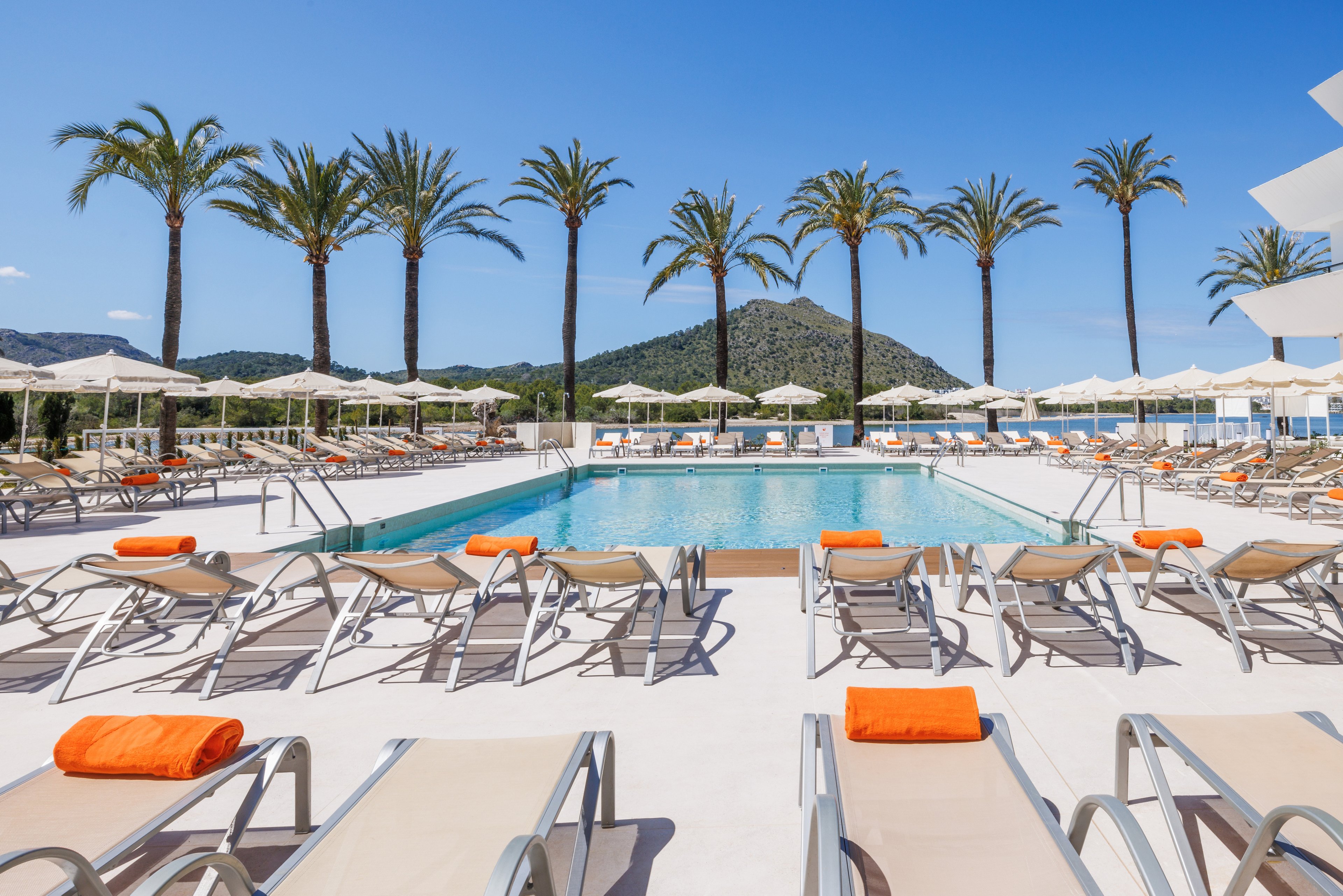 An outdoor resort swimming pool is surrounded by rows of empty sun loungers and umbrellas under a clear blue sky. Tall palm trees line the far side of the pool, with green hills and a mountain in the background. Bright orange towels on the loungers add a vivid contrast to the neutral deck and furniture. The scene suggests a calm, sunny vacation atmosphere at a modern holiday resort.