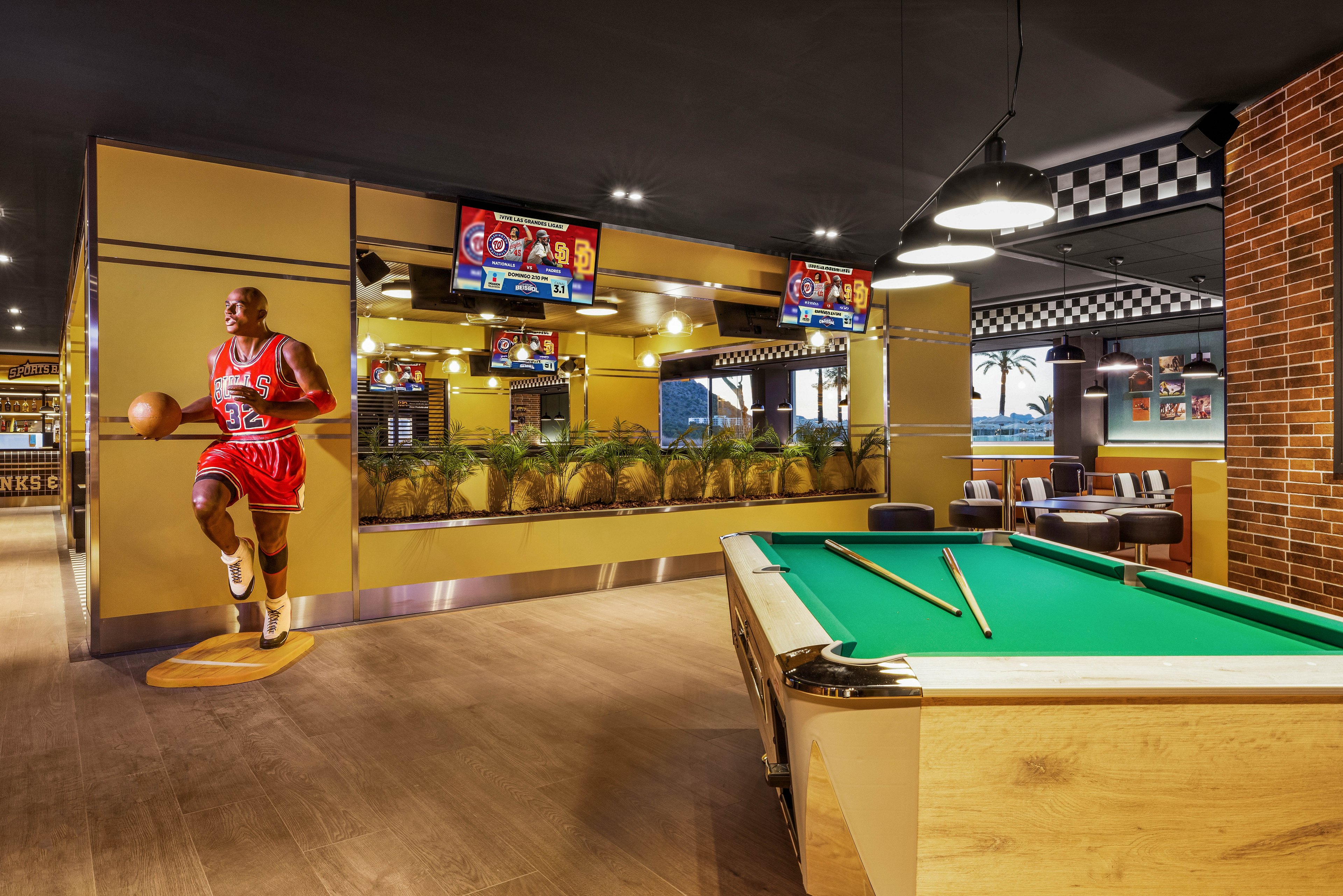 A modern sports bar features a green-felt pool table in the foreground under warm ambient lighting. In the background, a life-size basketball player cutout stands near a mirrored wall and indoor plants. Multiple flat-screen televisions display a sports broadcast above the seating area. The space combines wood, brick, and checkered design elements for a casual, entertainment-focused atmosphere.