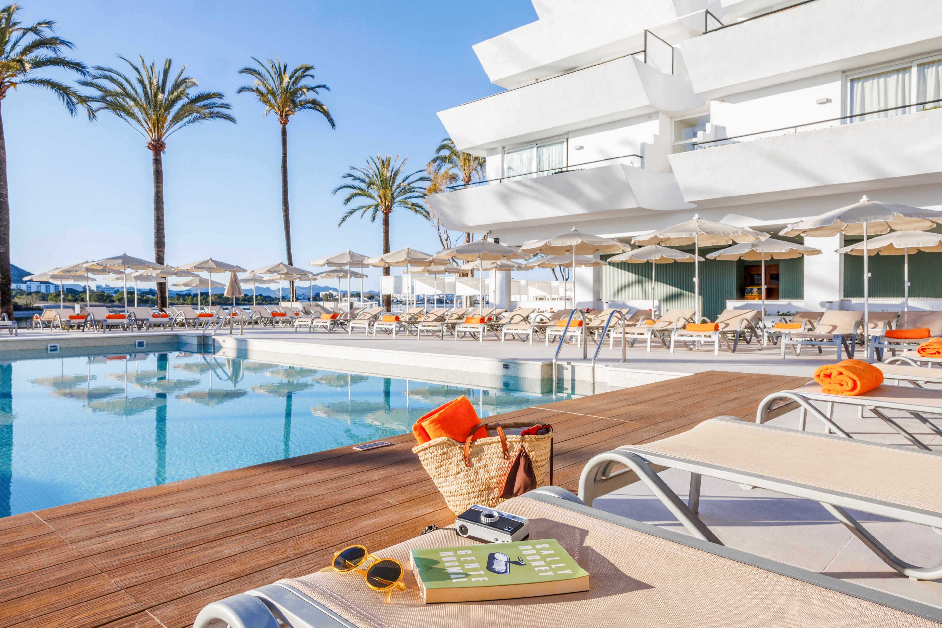 A bright resort swimming pool is surrounded by sun loungers and beige umbrellas on a clear day. In the foreground, a wicker beach bag, towel, sunglasses, and camera rest on a lounge chair beside the water. The modern white hotel building rises behind the pool, with balconies overlooking the scene. Tall palm trees and a blue sky enhance the relaxed, vacation atmosphere.