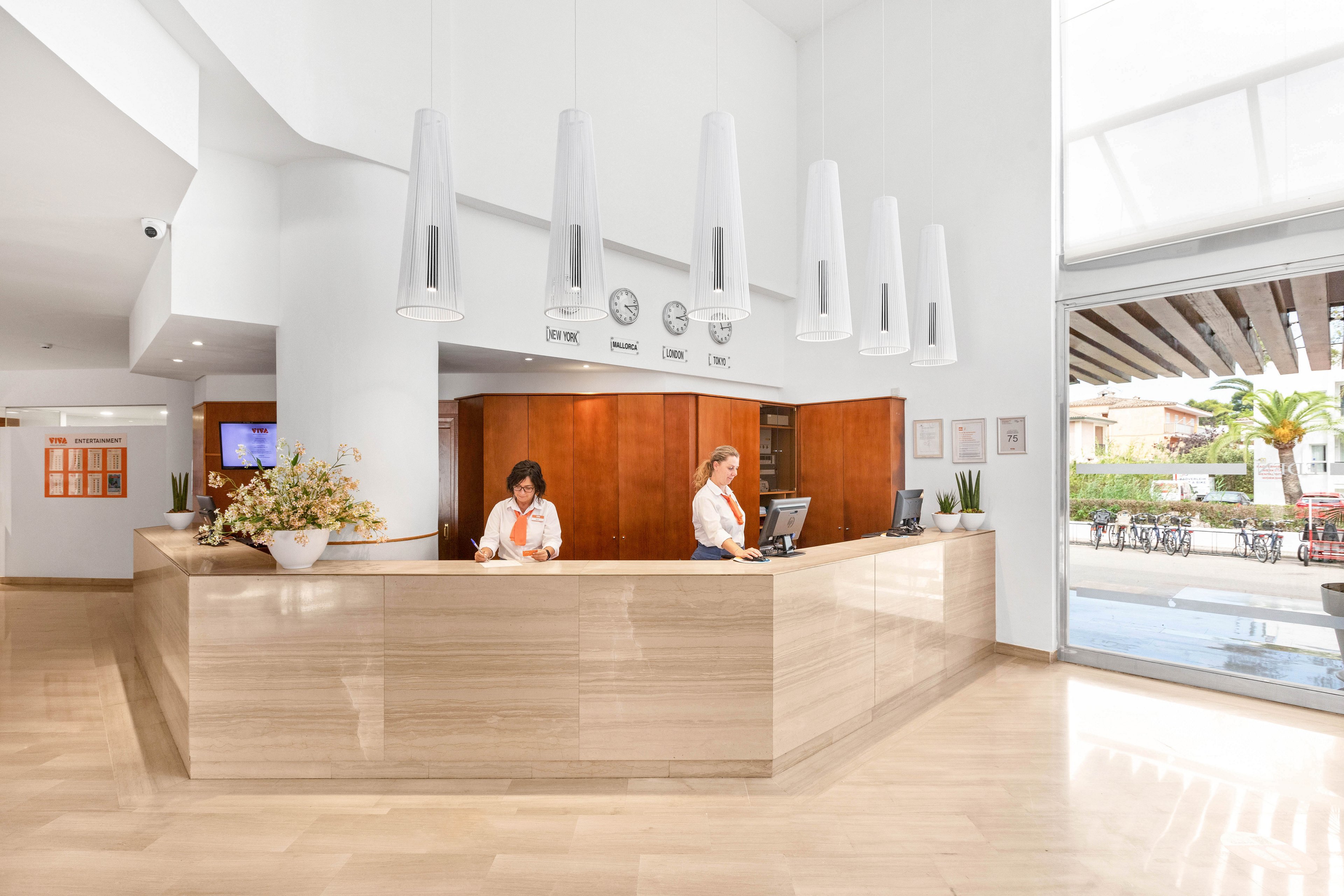 A spacious, modern hotel reception desk is staffed by two employees in coordinated uniforms. The lobby features light wood finishes, white walls, and contemporary pendant lights hanging above the counter. Several clocks are mounted on the wall behind the desk, and decorative plants and flowers add warmth to the space. Large windows and glass doors open to an outdoor seating area with tables and chairs, allowing natural light to fill the interior.