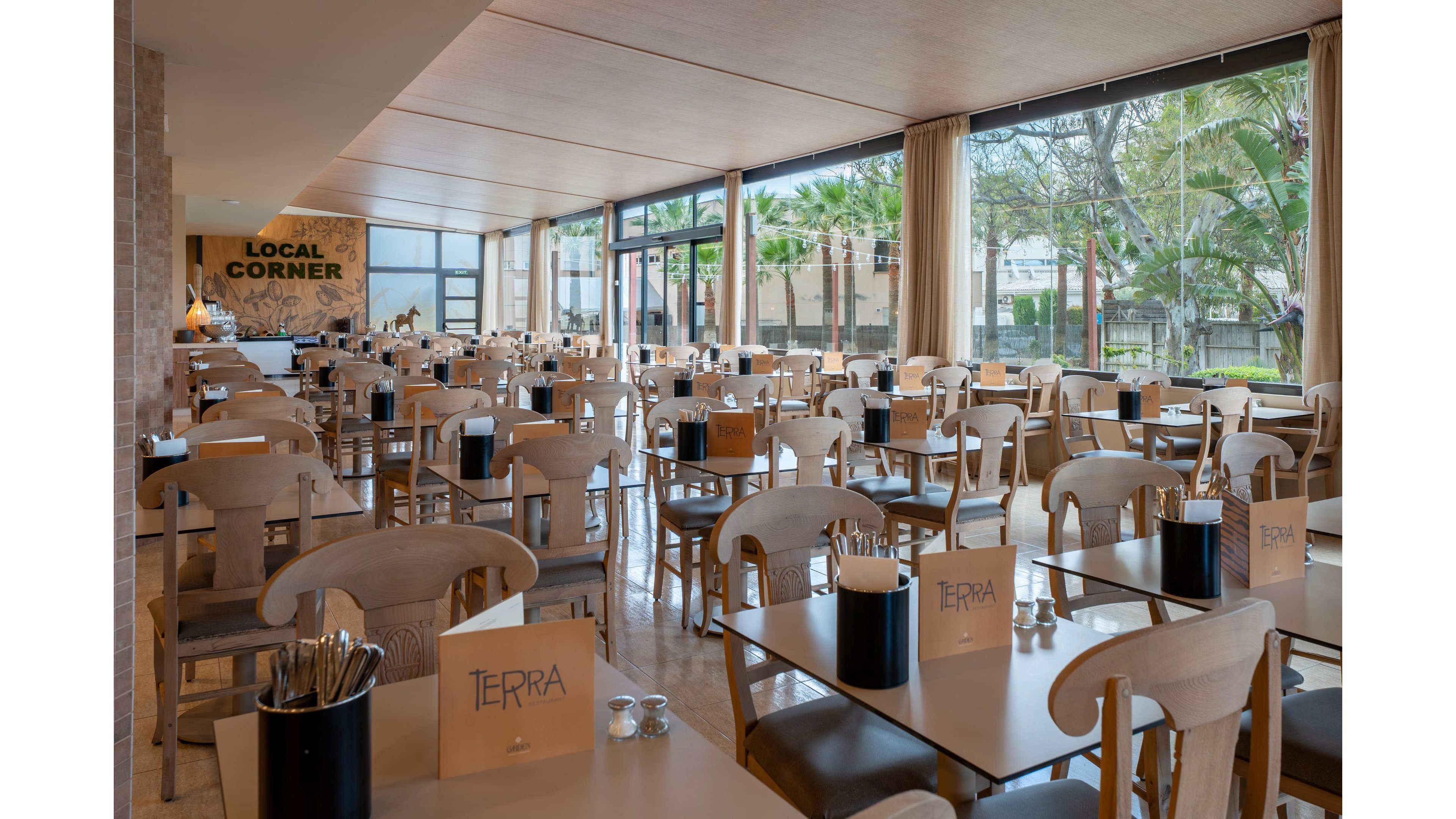 Spacious restaurant dining area with neatly arranged wooden tables and chairs stands empty, ready for guests. Large floor-to-ceiling windows line one side, revealing palm trees and greenery outside and filling the room with natural light. Table settings include menus labeled "TERRA" and containers holding cutlery and napkins. A sign reading "LOCAL CORNER" is visible near the buffet or service area at the back of the room.