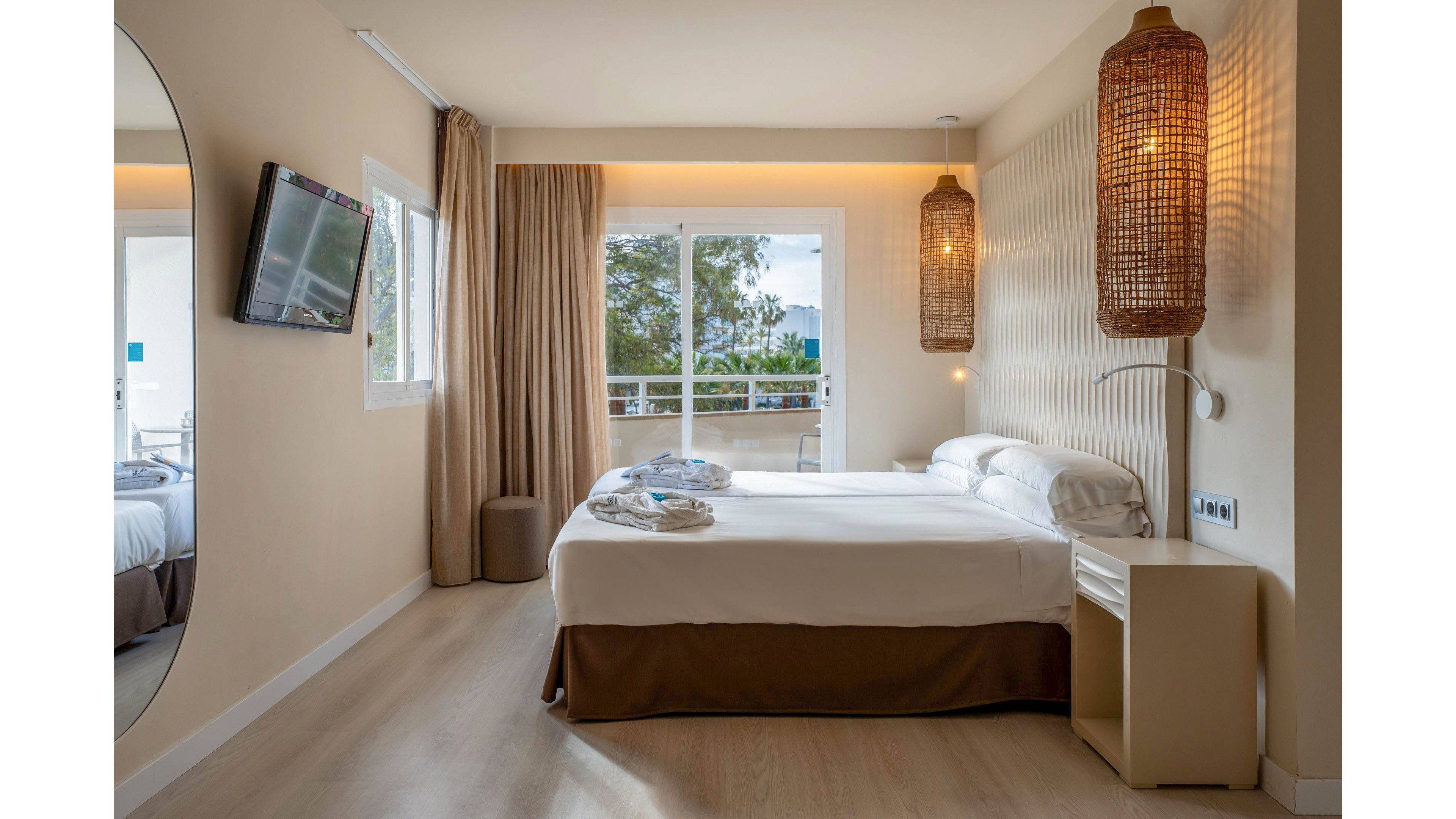 A contemporary hotel bedroom features a neatly made double bed with white linens and a brown bed skirt. Large sliding glass doors open to a balcony with a view of trees and distant hills. Warm beige walls, wicker pendant lights, and floor-length curtains create a cozy, inviting atmosphere. A wall-mounted television and minimalist bedside table complete the clean, modern design.