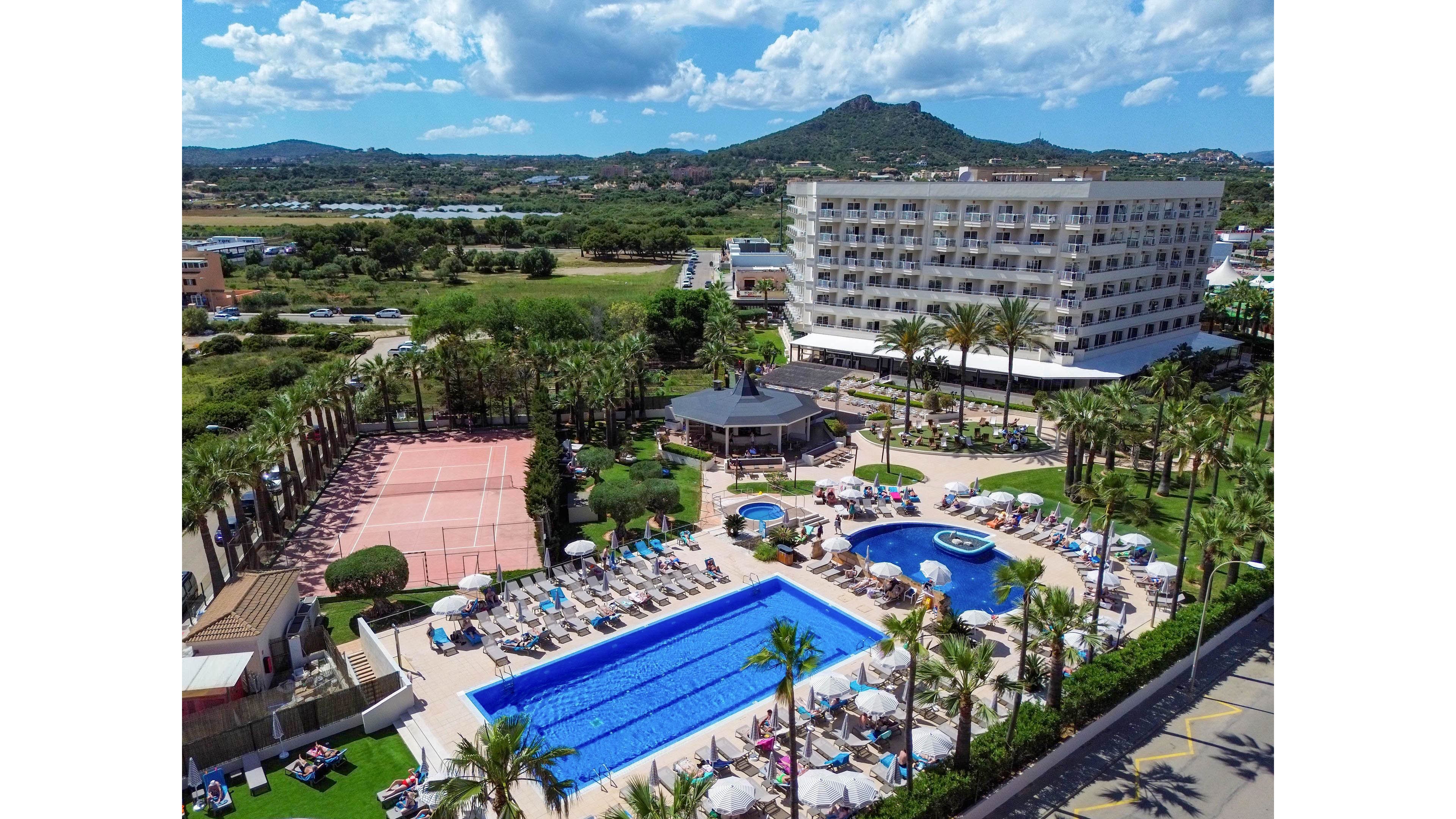 A large resort hotel complex is shown from an elevated aerial perspective, featuring a central rectangular swimming pool surrounded by sun loungers and umbrellas. Adjacent to the main pool is a smaller circular pool and landscaped areas with palm trees. To the side, there are multiple tennis courts and additional recreational spaces. In the background, open green fields and distant hills extend under a bright blue sky with scattered clouds, creating a relaxed vacation atmosphere.