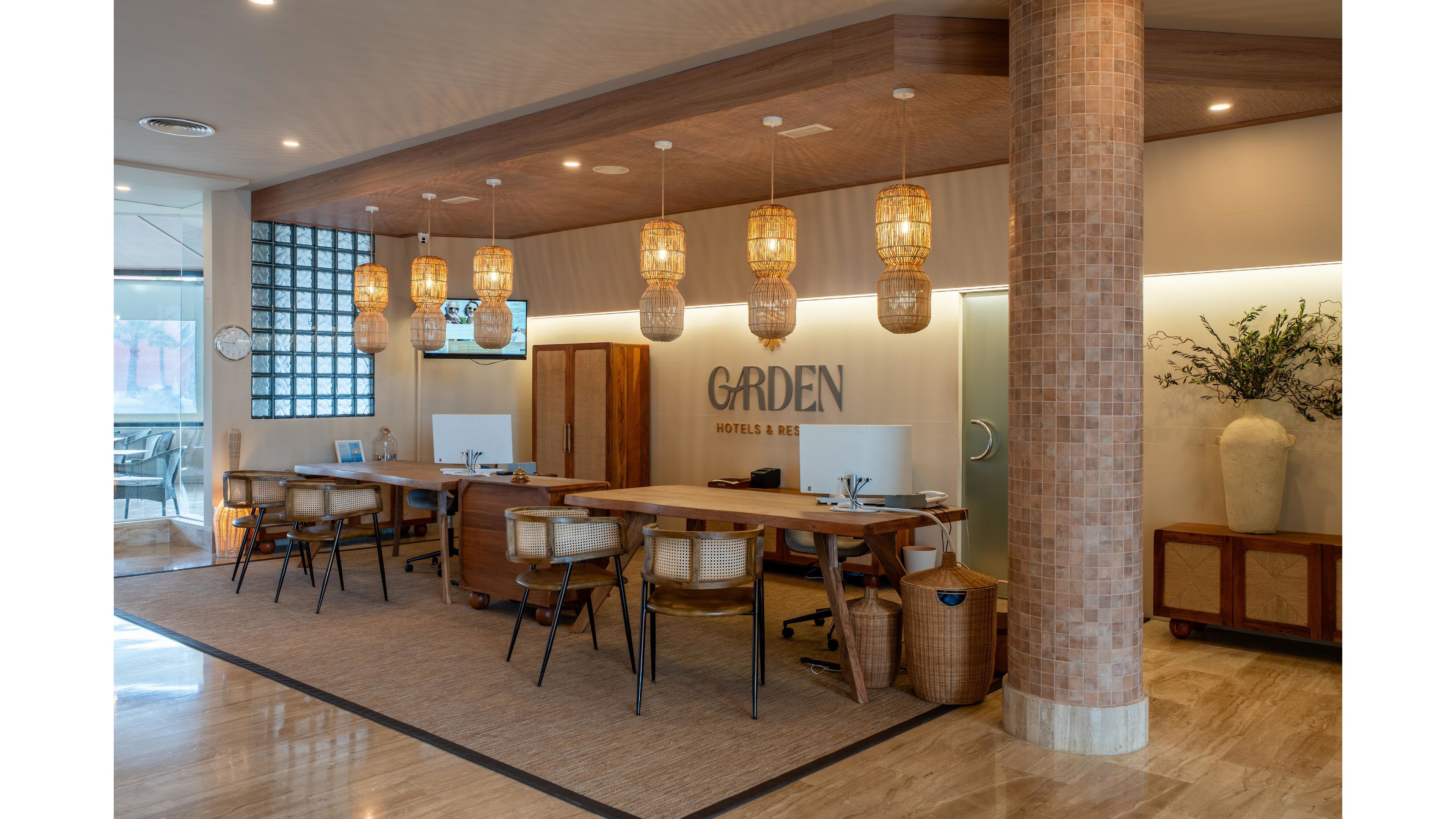 Contemporary office workstations are arranged in a row beneath hanging woven pendant lights. The space features wooden desks, rattan chairs, and a tiled column that complement the neutral flooring. A glass wall and door add openness, while plants and decorative objects soften the interior. The word "GARDEN" appears prominently on the back wall, suggesting the name of the space.