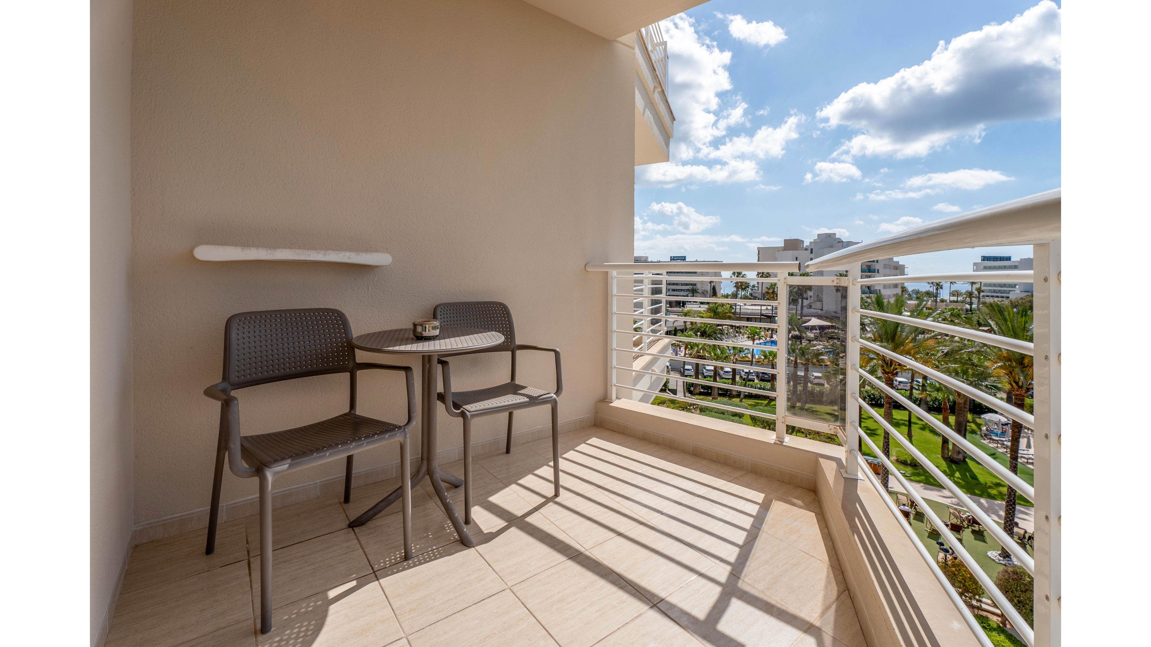 A small hotel balcony features two modern chairs and a compact table in bright daylight. The tiled floor and light-colored walls reflect the sun, creating strong shadows across the space. A metal railing frames a view of palm trees, buildings, and a distant body of water under a partly cloudy sky. The scene suggests a relaxing outdoor spot for guests to sit and enjoy the warm weather and scenery.