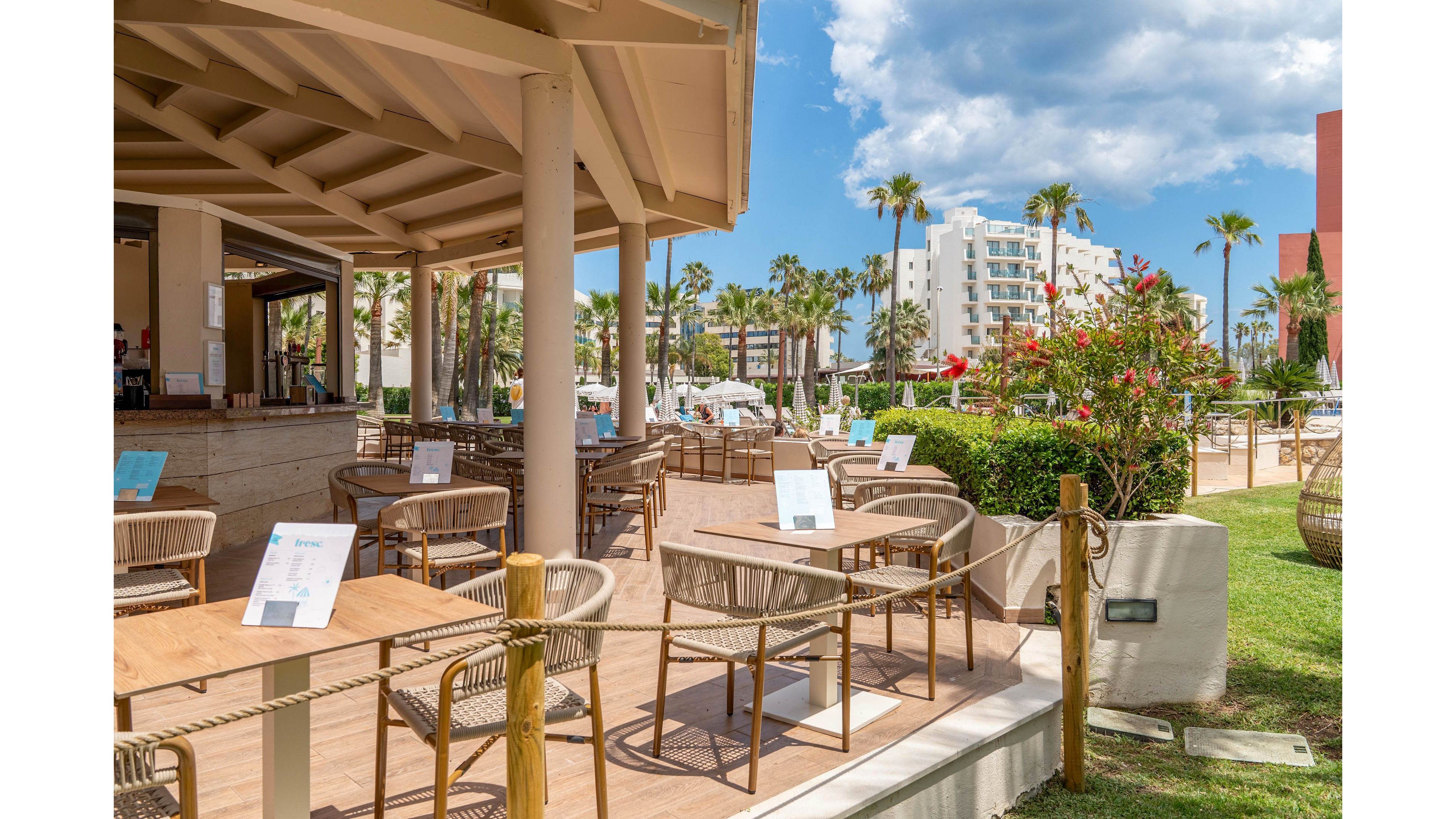An open-air café terrace at a resort features neatly arranged wooden tables and wicker chairs. Menus are placed on each table, and the area is bordered by rope barriers and wooden posts. In the background, guests relax near a swimming pool surrounded by palm trees and sun loungers. Modern white resort buildings and a partly cloudy blue sky complete the sunny vacation atmosphere.
