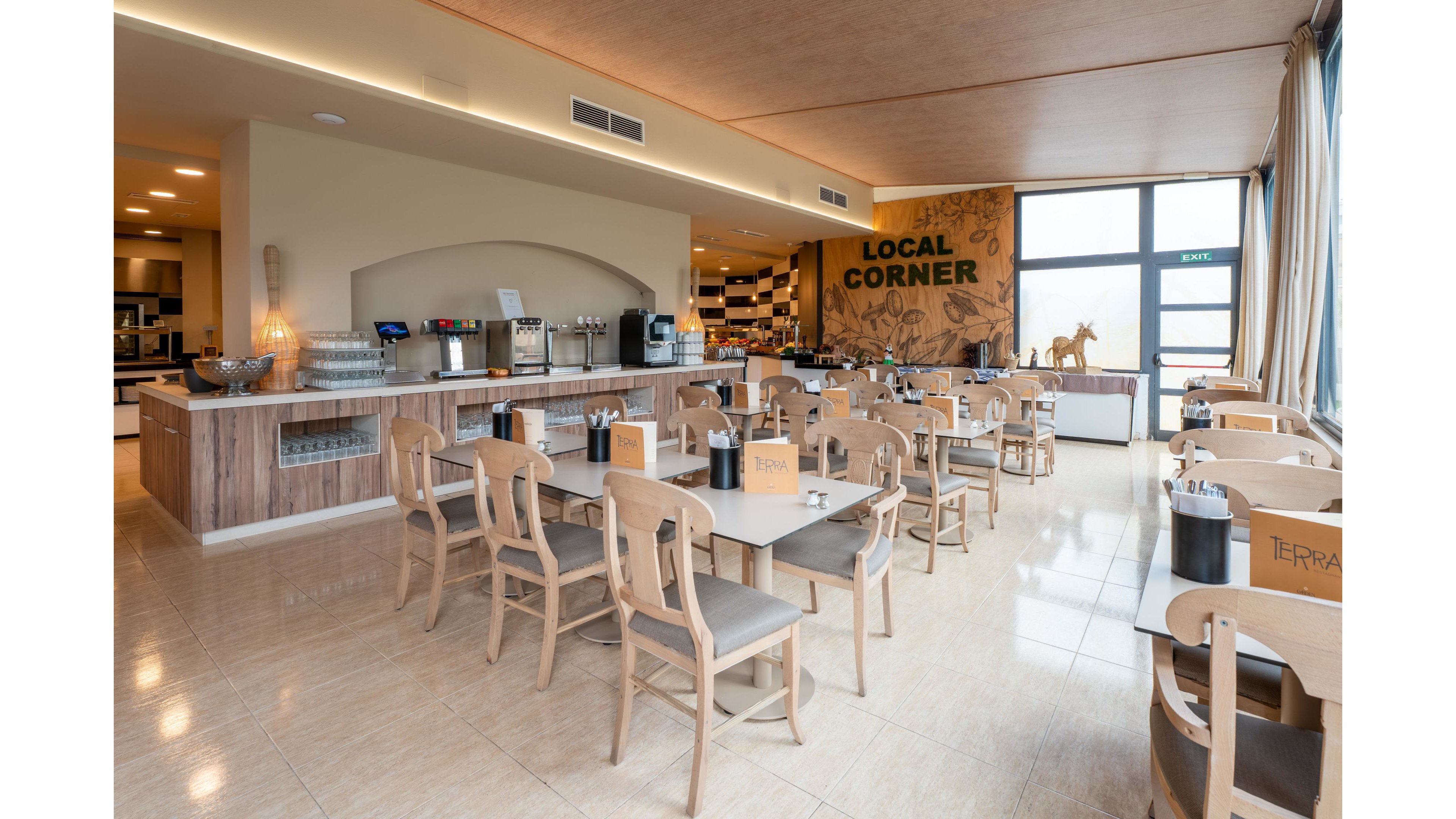 A spacious, modern cafeteria interior features light wooden tables and chairs neatly arranged in rows. The serving counter in the background displays drink dispensers, cups, and other self-service items. A large wall graphic reading "LOCAL CORNER" decorates the far wall beside tall windows that let in natural light. The space appears clean, empty of people, and ready for guests, with neutral tones and contemporary decor.