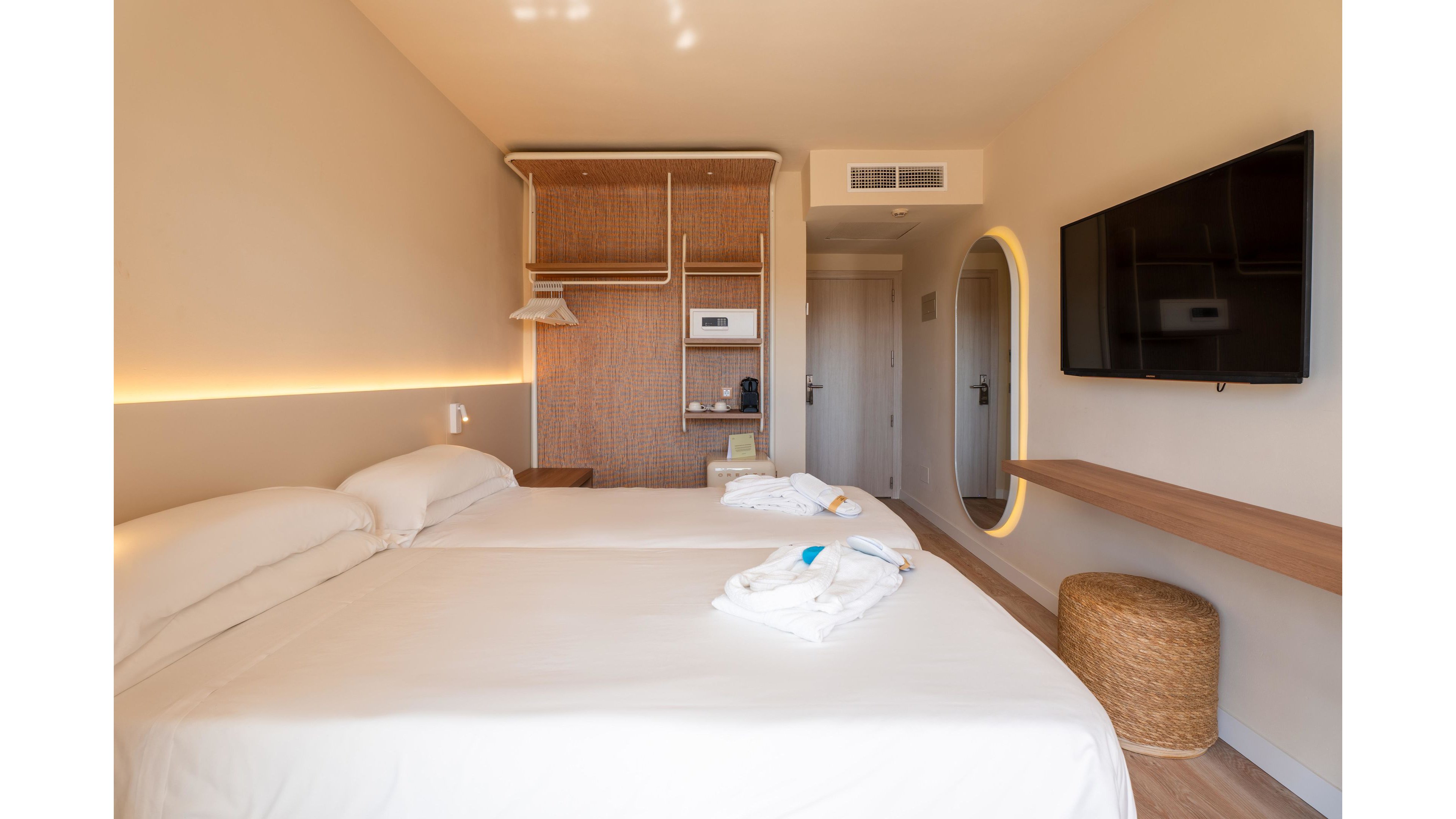 A bright modern hotel room features two neatly made twin beds with white linens. The space includes a wall-mounted flat-screen TV opposite the beds and a floating wooden shelf with a woven stool beneath it. At the far end, built-in wooden shelving, a small wardrobe, and a coffee station are visible near the entrance door. Warm neutral tones, indirect lighting, and minimal decor create a calm, contemporary atmosphere.