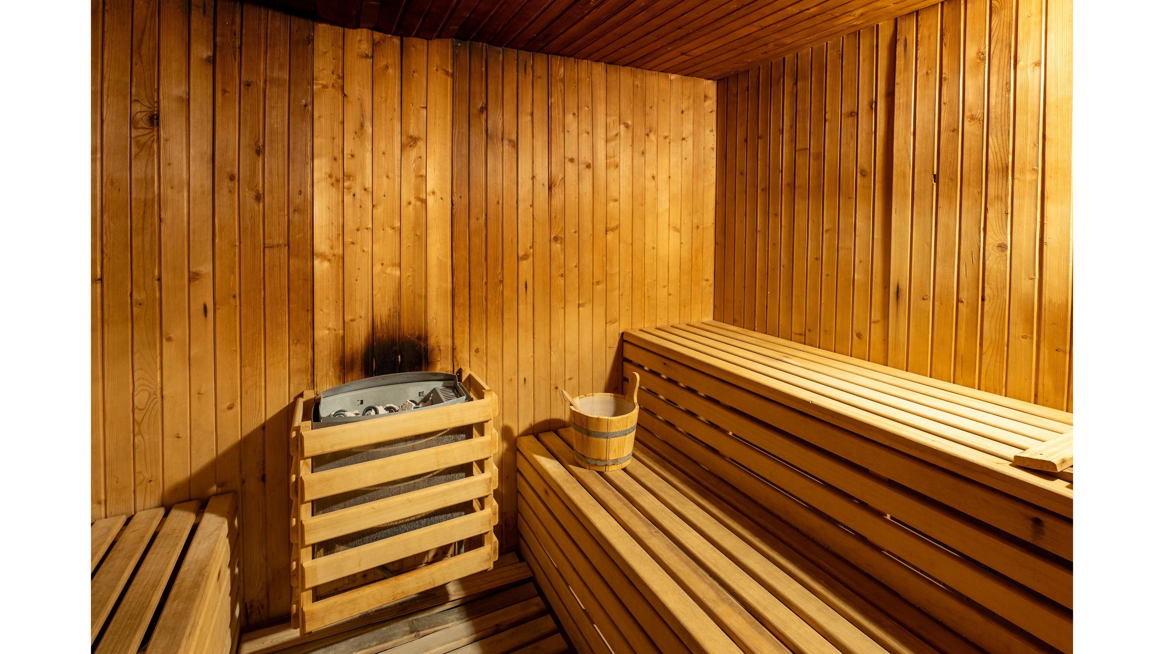 A traditional wooden sauna interior is shown with tiered benches along the walls. A metal sauna heater filled with stones is enclosed in a wooden guard on the left side. A wooden bucket and ladle rest on one of the benches, ready for use. Warm lighting enhances the golden tones of the wood, creating a cozy and relaxing atmosphere.