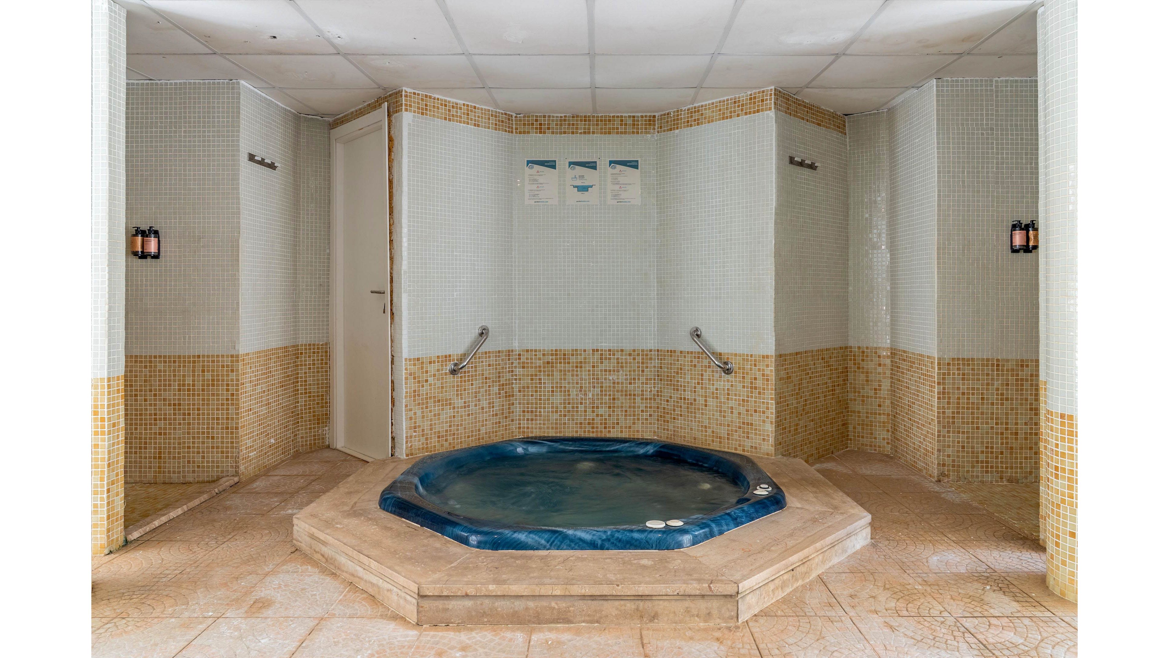 An empty blue jacuzzi tub is set into a raised tiled platform in an indoor spa room. The surrounding walls are covered with small mosaic tiles in white and beige tones. Metal handrails lead into the tub, and wall-mounted lights provide subtle illumination. A closed door and a small set of wall notices are visible in the background, emphasizing a quiet, private wellness space.