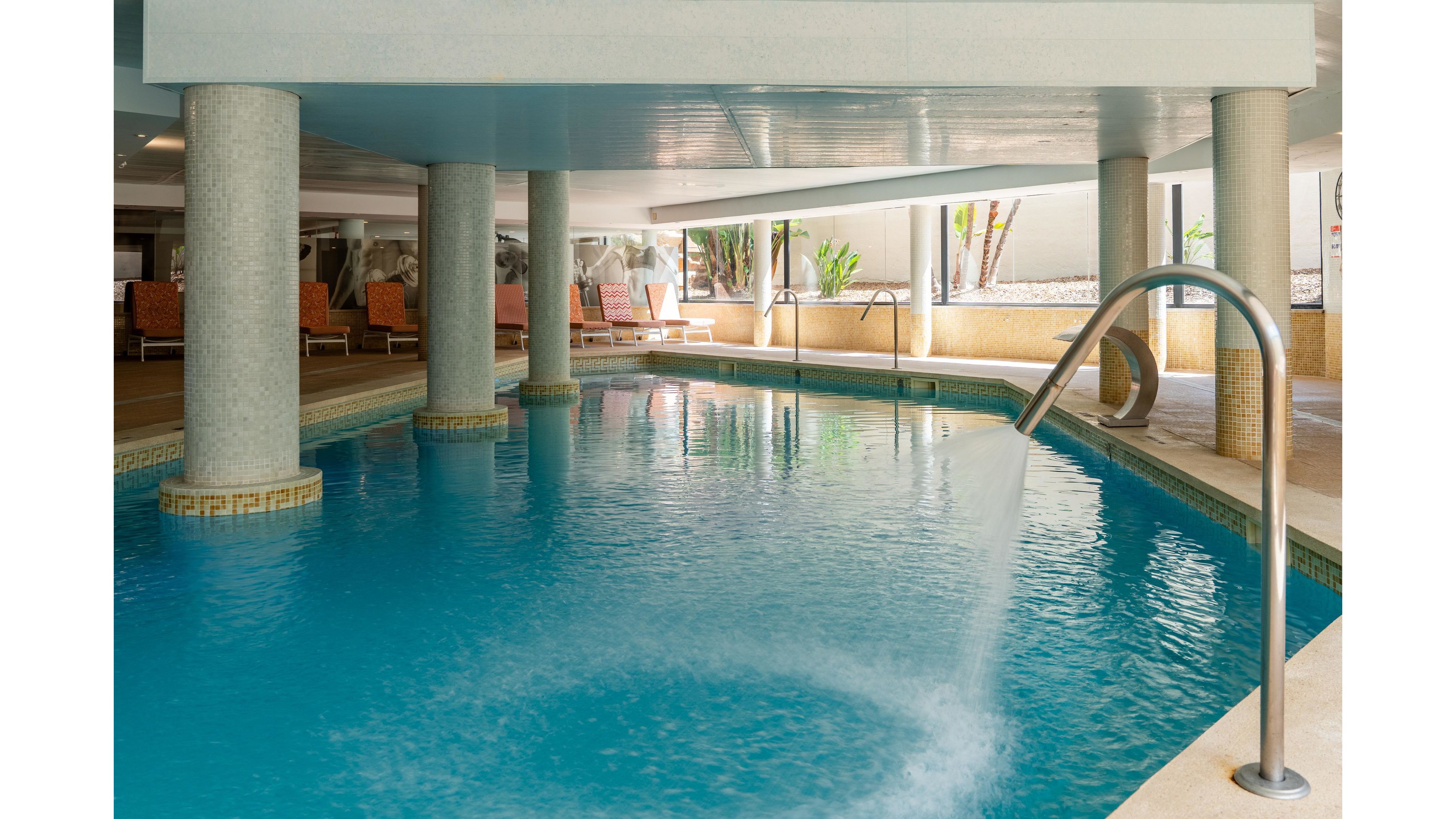 An indoor swimming pool features clear blue water and a curved metal fountain spout creating a gentle spray. The pool is surrounded by sturdy columns and a tiled deck area. In the background, cushioned lounge chairs and potted plants suggest a relaxing hotel or spa environment. Natural light filters in from large openings, adding a calm and inviting atmosphere.