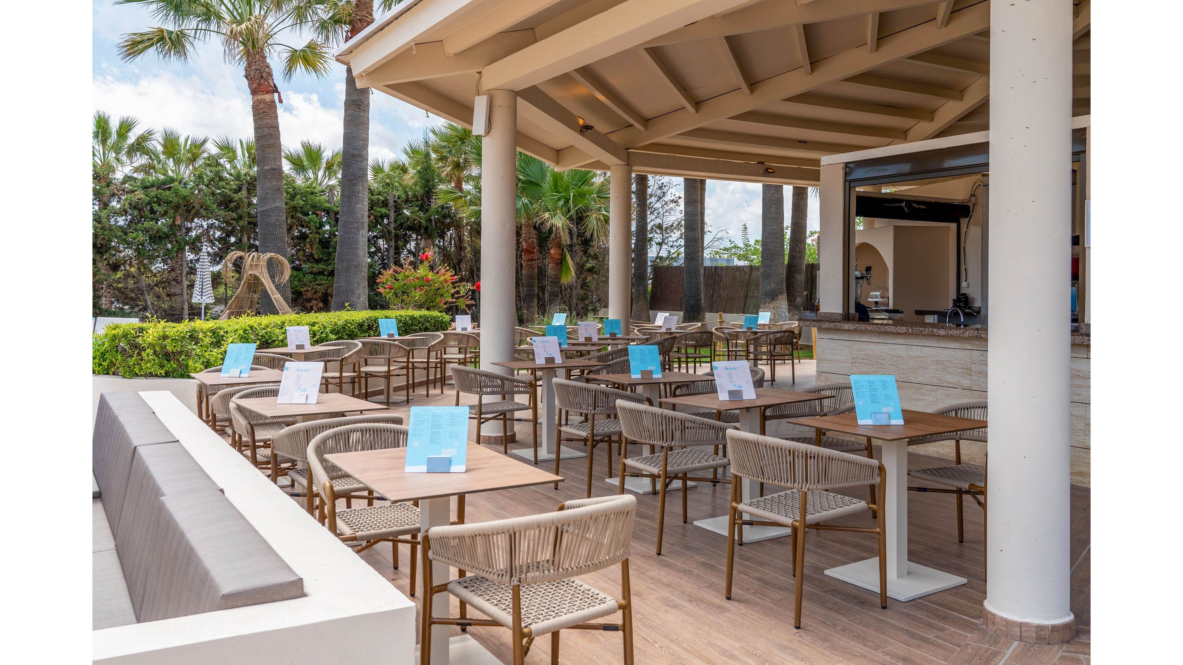 An open-air café terrace features neatly arranged wooden tables and wicker chairs under a covered patio. Each table has a blue menu card standing upright, while the bar counter is visible in the background. Palm trees, lush greenery, and a bright sky create a relaxed resort atmosphere. The seating area appears ready for guests but is currently empty, emphasizing calm and spaciousness.