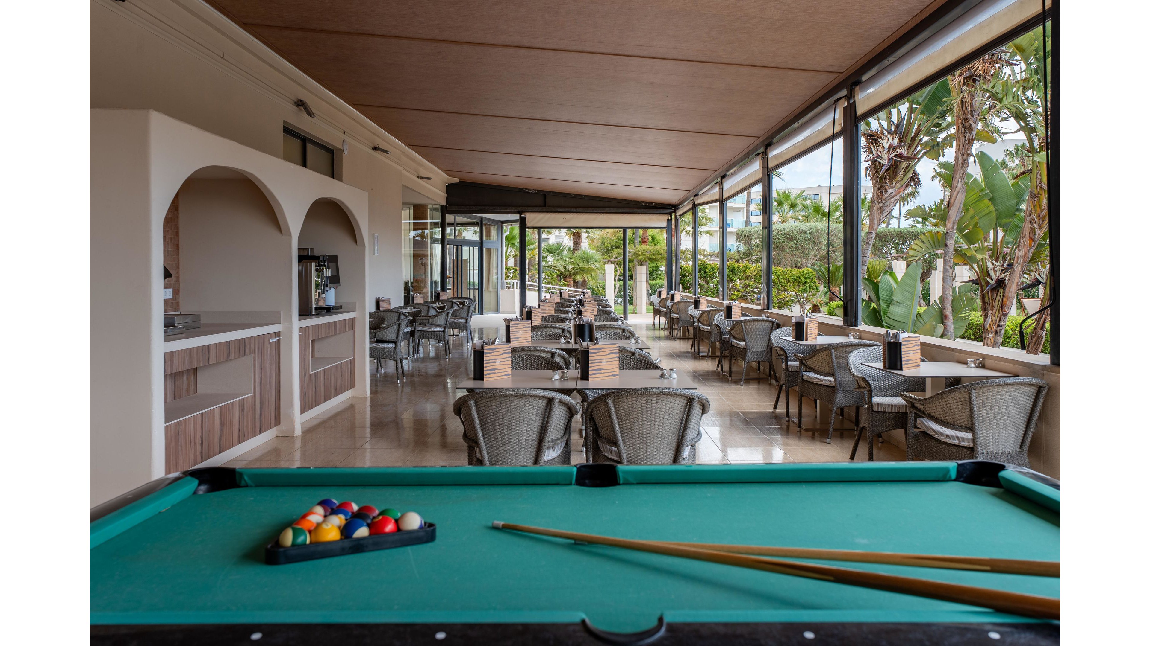An open-air resort terrace features a green pool table in the foreground with racked billiard balls and two wooden cues. Beyond the table, rows of wicker chairs and small tables line the covered patio, creating a relaxed lounge and dining area. Large windows and open sides reveal lush tropical plants and palm trees outside. The space appears calm and inviting, suitable for leisure, socializing, and casual recreation.