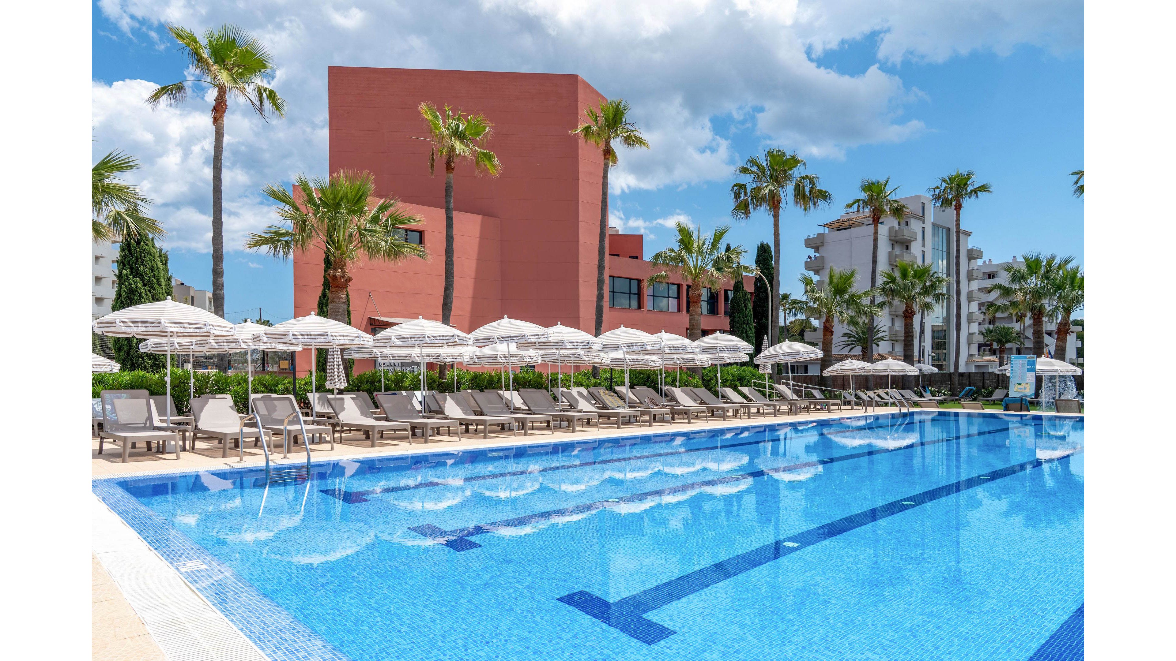 A large outdoor swimming pool stretches alongside rows of sun loungers and white umbrellas at a modern resort. Tall palm trees line the pool area, adding a tropical feel to the scene. In the background, contemporary buildings with red and white facades rise against a bright blue sky with scattered clouds. The clear, calm water and empty loungers suggest a peaceful, relaxing vacation setting.