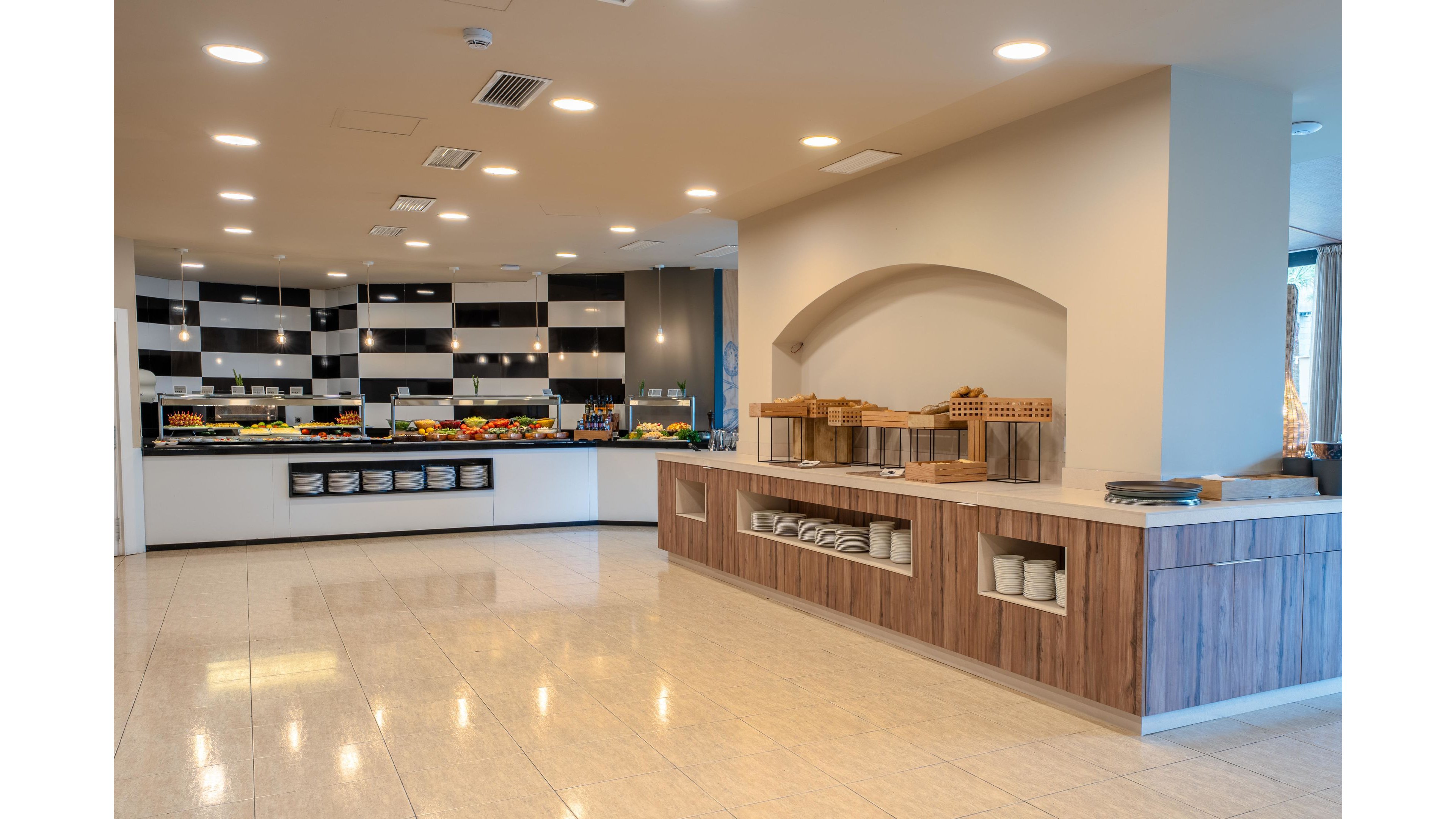 A spacious self-service buffet area is shown in a modern dining facility. Long wooden and white counters hold empty plates, baskets, and serving stations ready for food service. The back wall features a black and white tiled design with neatly arranged dishes and colorful food displays. Bright ceiling lights and clean tiled floors create an inviting, contemporary atmosphere.