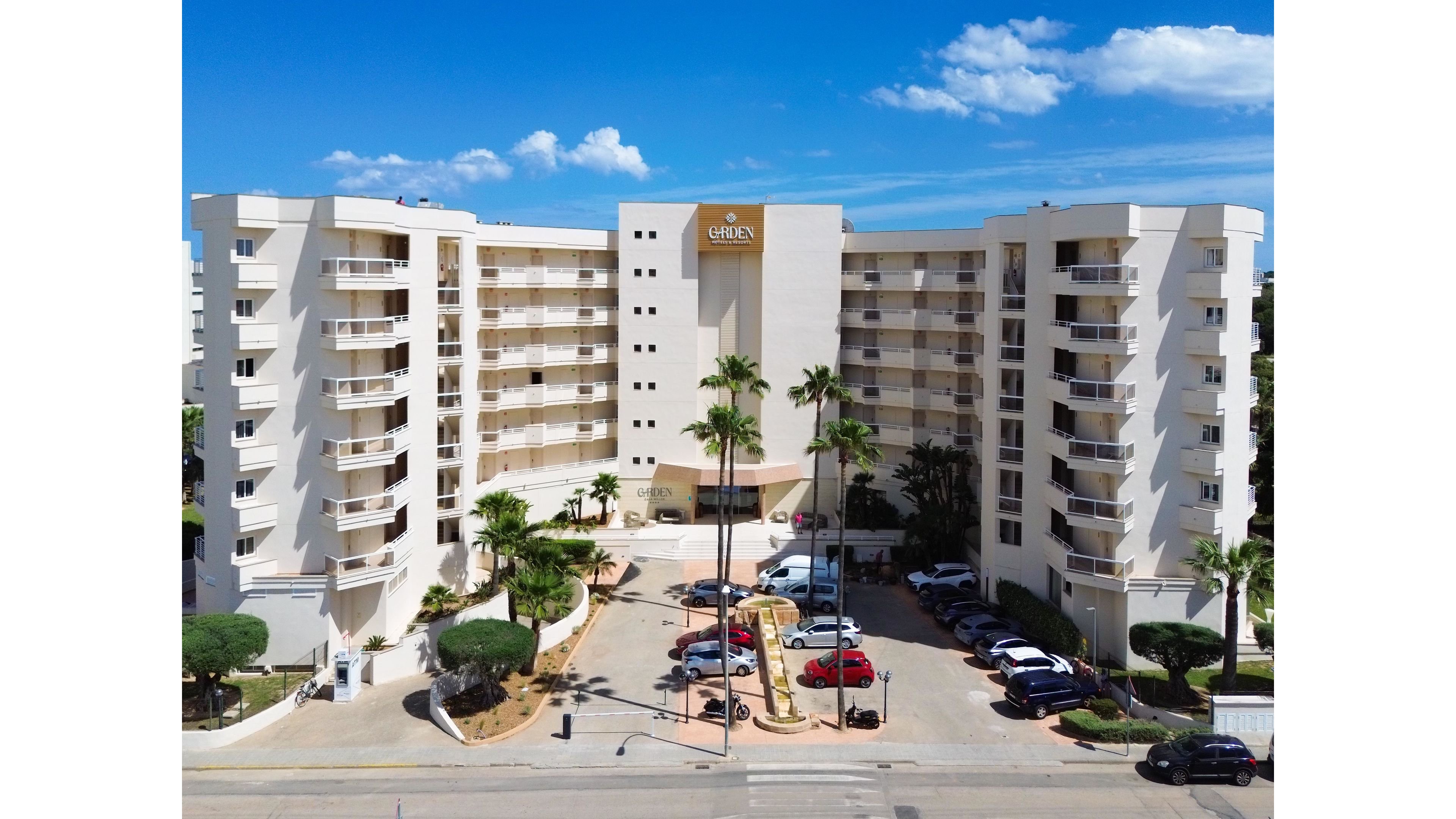 A large beachfront resort hotel stands prominently with two symmetrical wings and multiple balconies facing the street. The building features a central entrance canopy, landscaped areas with trimmed trees, and tall palm trees lining the driveway. Several parked cars are visible in front of the hotel, suggesting guest arrivals and departures. The scene is captured on a bright, sunny day with a clear blue sky and scattered clouds above.
