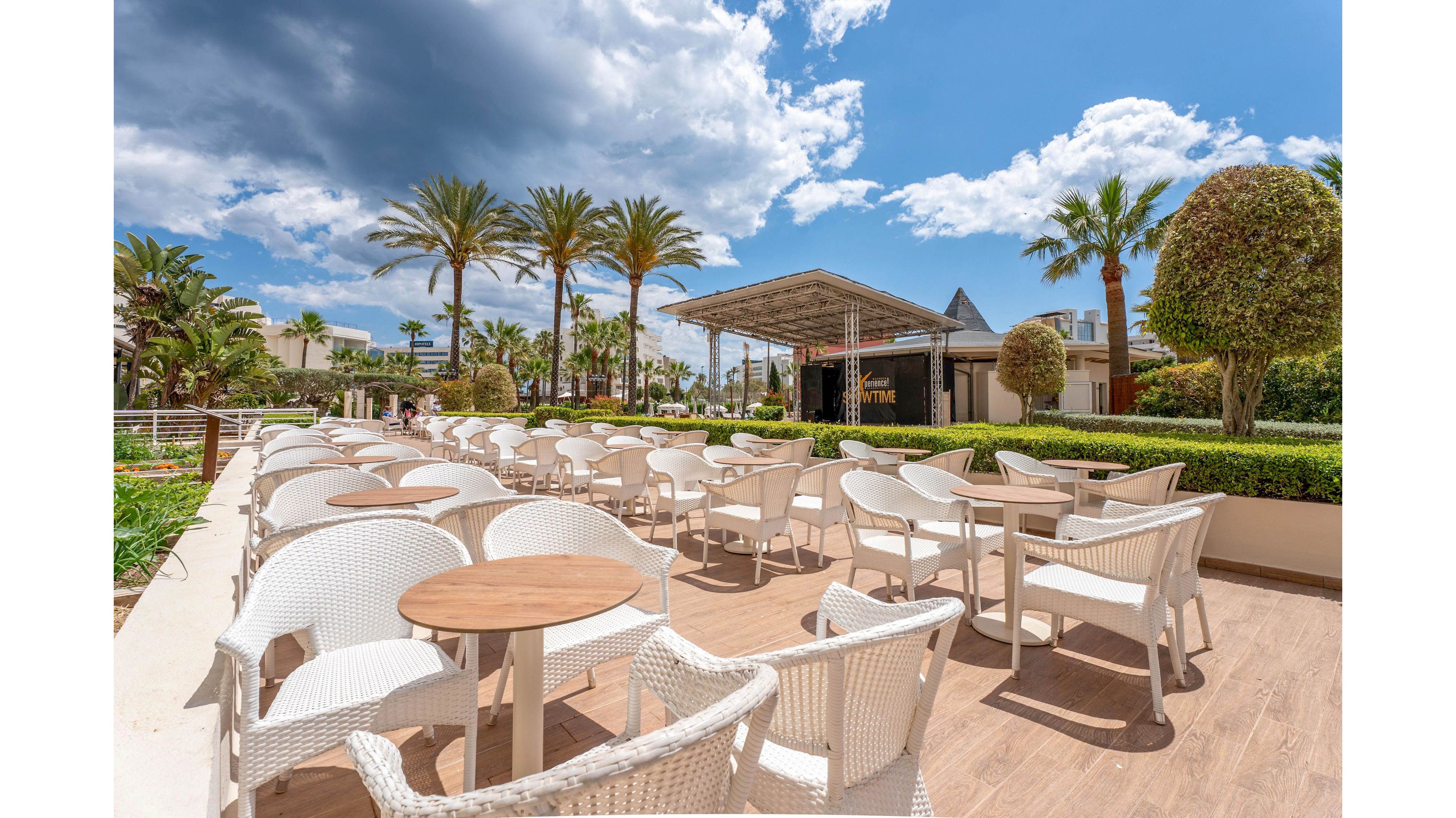 An empty outdoor terrace is arranged with rows of white wicker chairs and round wooden tables. The seating area is part of a resort or hotel, bordered by manicured hedges and sculpted trees. Tall palm trees line the background, leading toward a covered stage or pavilion structure. A bright blue sky with dramatic clouds adds contrast and depth to the sunny scene.