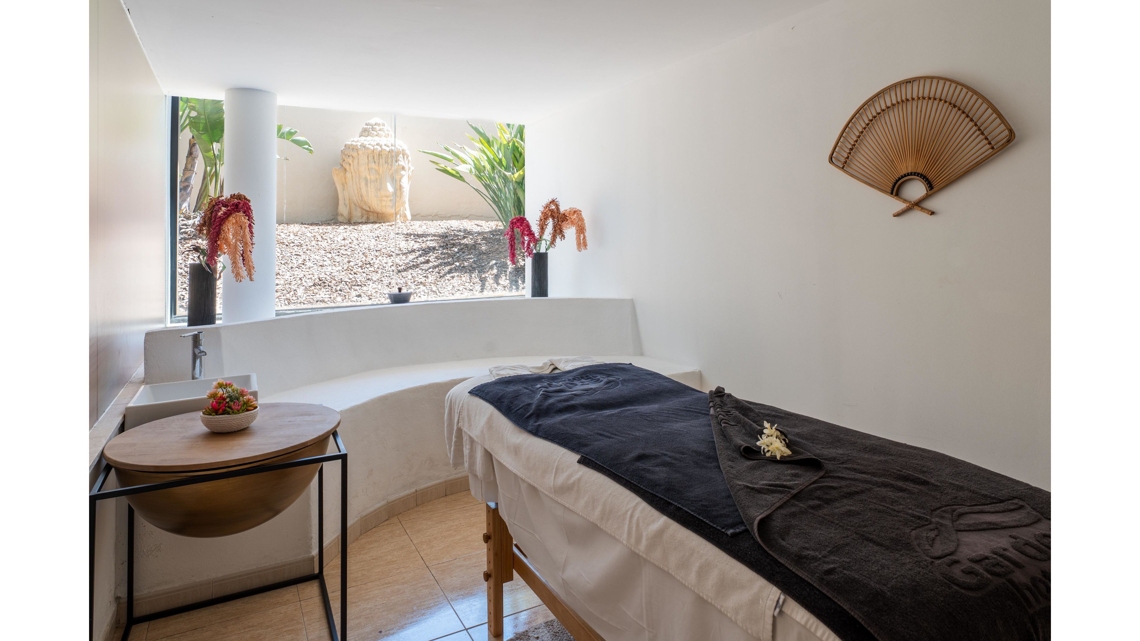 A tranquil spa massage room features a treatment table covered with white linens and a dark blanket. The space is brightened by natural light from a wide window overlooking a minimalist rock garden and plants. A small side table holds a decorative bowl, while wall-mounted fans and hanging dried flowers add a touch of warmth. The clean, neutral walls and tiled floor create a calm, relaxing atmosphere ideal for wellness treatments.