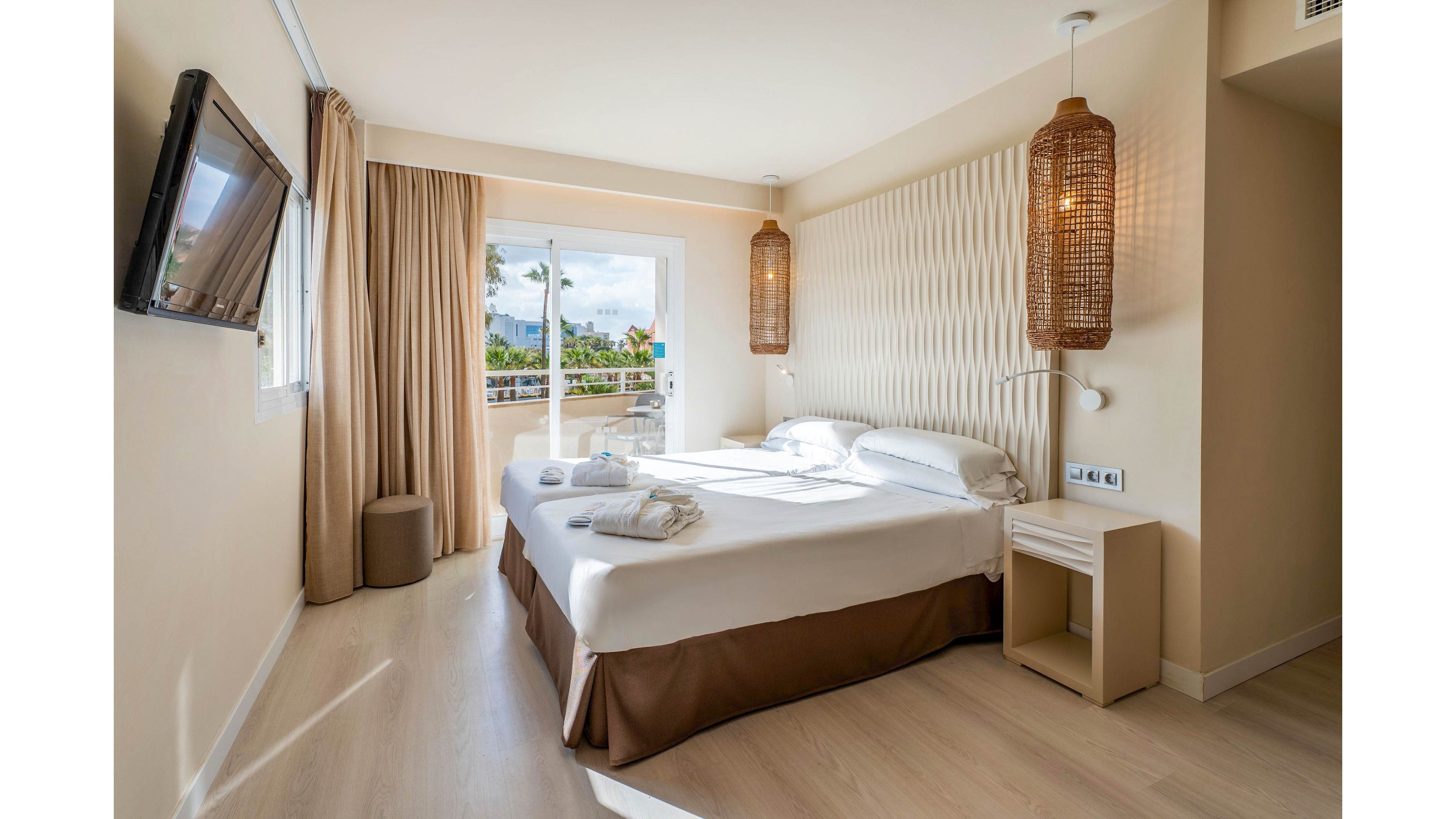 A bright, modern hotel bedroom features a neatly made double bed with white linens and folded towels. Natural light floods in through a glass door leading to a balcony with a partial outdoor view. Warm beige tones, wooden flooring, and woven pendant lamps create a cozy, contemporary atmosphere. A wall-mounted television and minimal bedside furniture complete the clean, inviting interior.