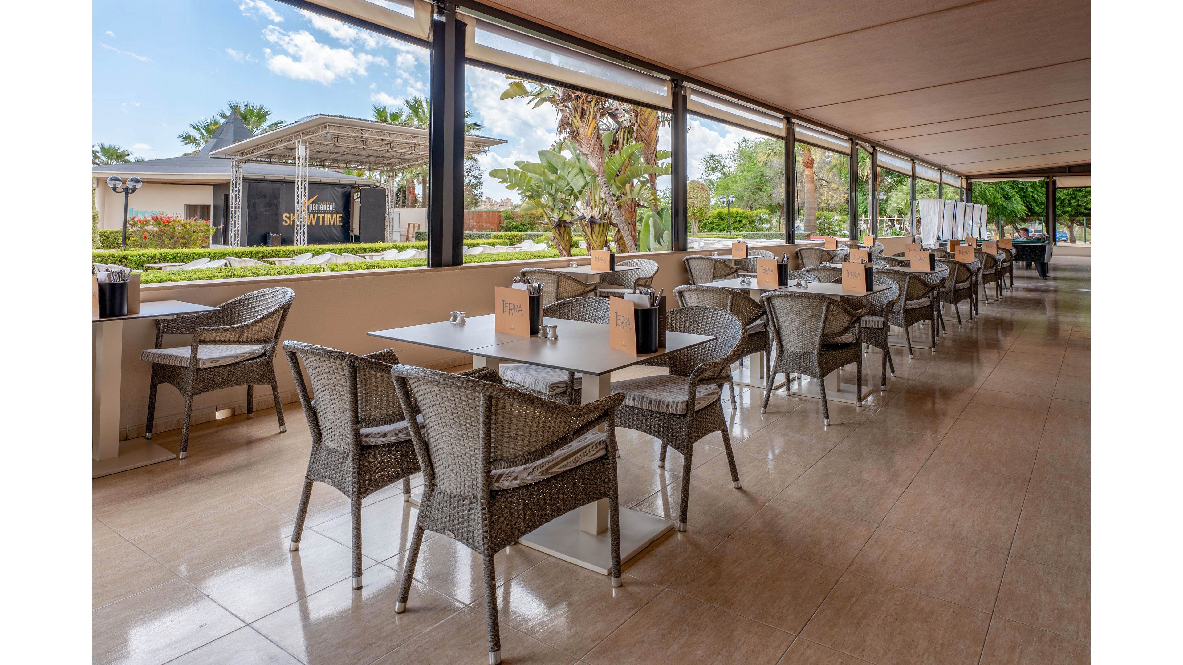 An open-air terrace dining area features rows of wicker chairs and square tables neatly arranged under a covered patio. The space overlooks a landscaped garden with palm trees and a stage structure visible in the background. Natural light floods the area through large open sides, creating an airy and relaxed atmosphere. Tabletop items and menus are placed on each table, suggesting a prepared but currently unoccupied restaurant setting.