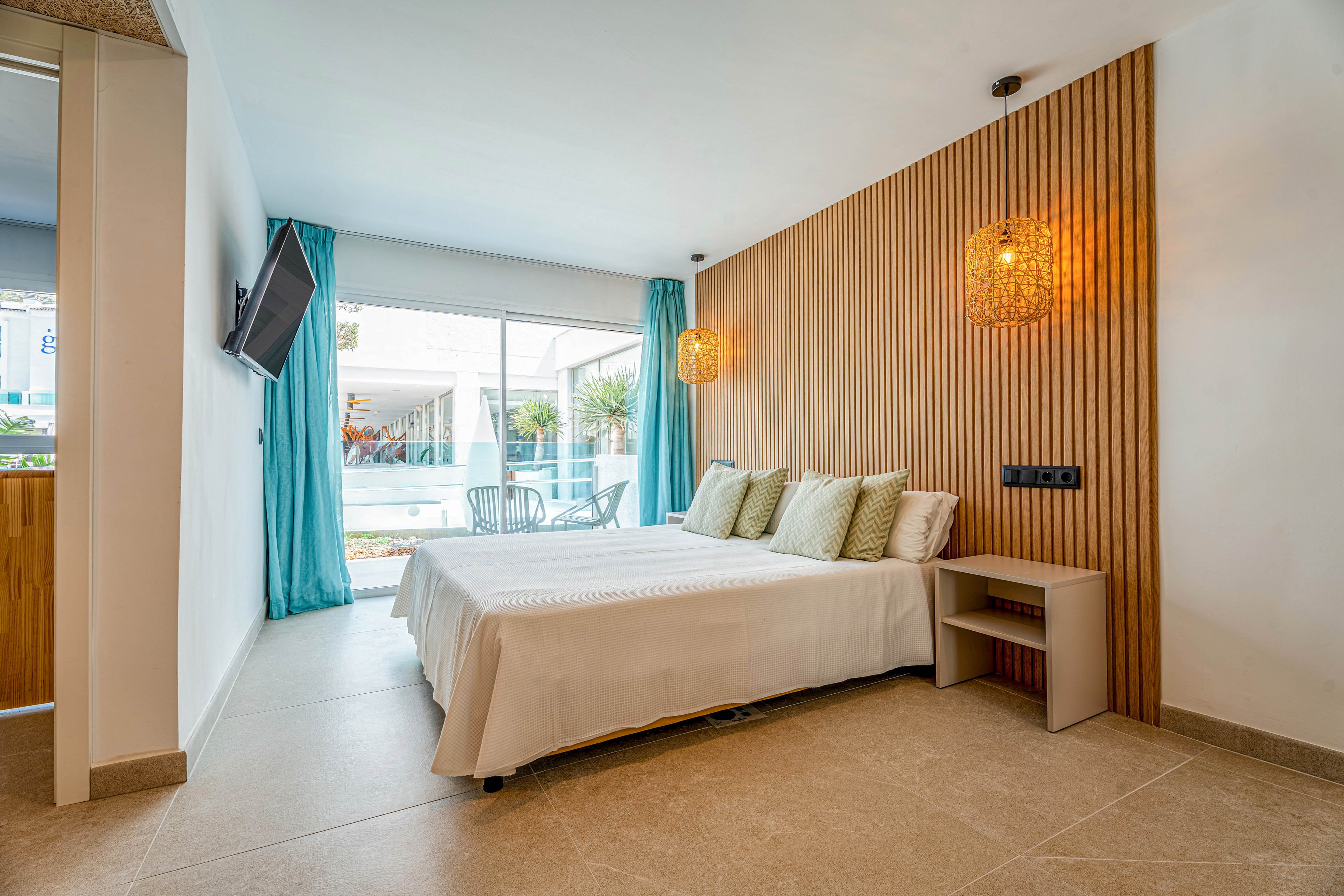 Bright modern hotel room featuring a neatly made double bed with multiple cushions. The space includes a wooden slat accent wall, hanging woven lamps, and a small bedside table. Large glass doors open to an outdoor terrace with chairs and greenery, allowing natural light to fill the room. Turquoise curtains and neutral flooring create a clean, relaxing atmosphere suitable for vacation or business travel.