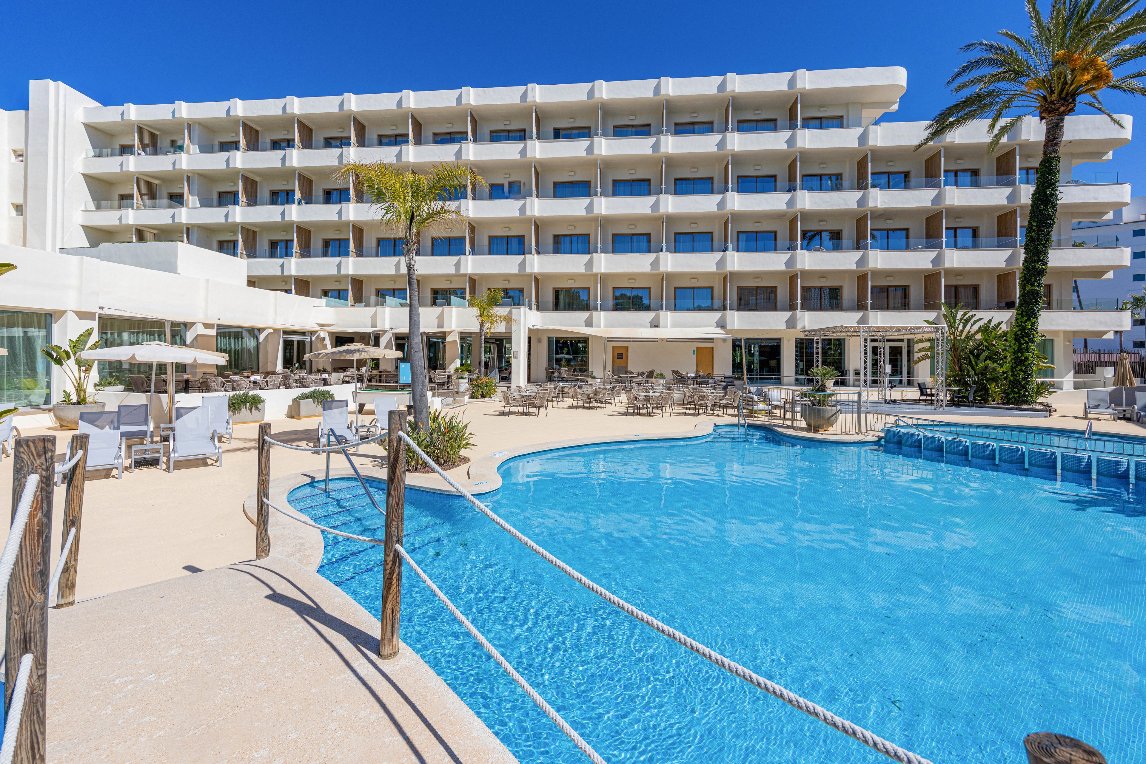 A contemporary multi-story hotel overlooks a large outdoor swimming pool under a clear blue sky. Sun loungers, umbrellas, and seating areas surround the pool, creating a relaxed resort atmosphere. Palm trees and minimalist white architecture emphasize a bright, coastal setting. The scene suggests a sunny vacation destination focused on leisure and relaxation.