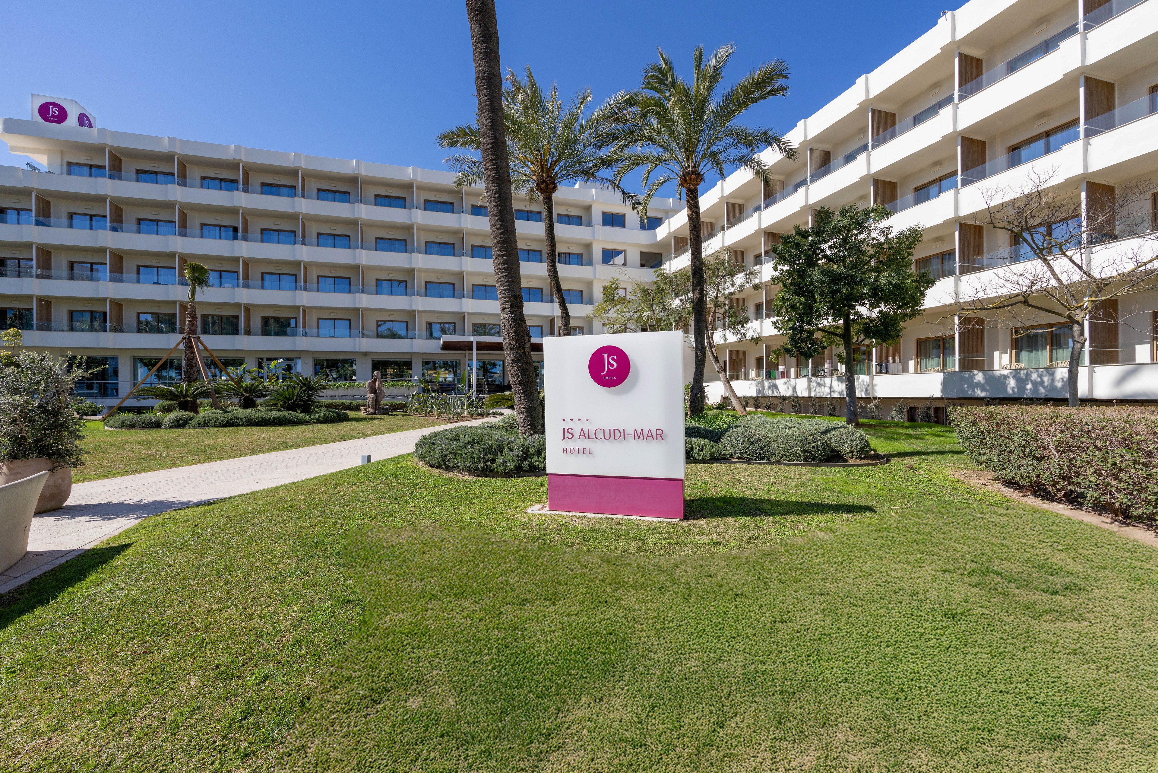 A modern multi-story hotel complex is shown with white balconies facing a neatly maintained green lawn. In the foreground, a large white and pink sign with the text "JS ALCUDI-MAR" stands on the grass. Palm trees and trimmed shrubs decorate the courtyard, creating a resort-like atmosphere. The clear blue sky and bright sunlight suggest a warm, coastal destination. No people are visible, emphasizing the architecture and outdoor space.