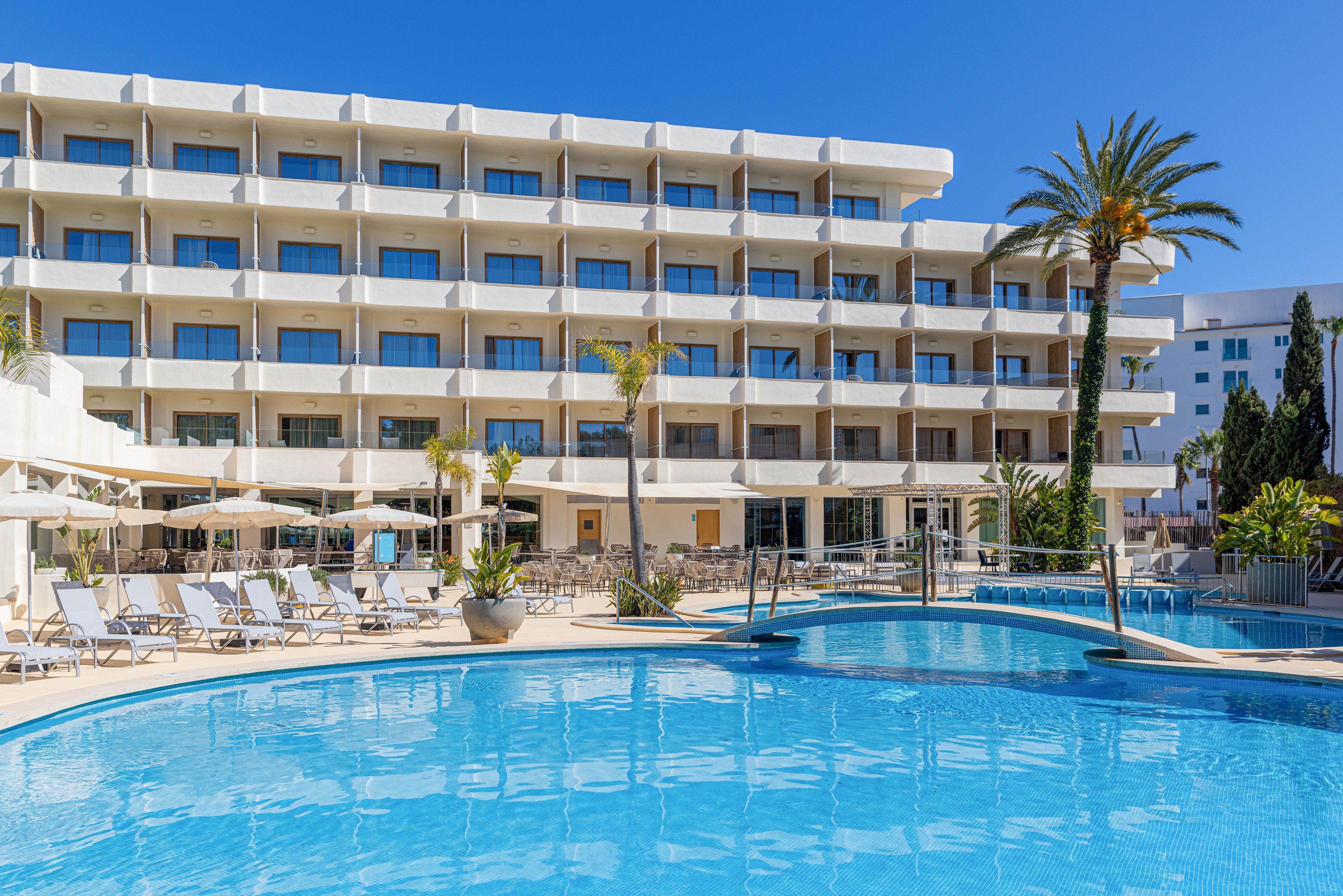 A contemporary multi-story hotel overlooks a large outdoor swimming pool. Sun loungers and umbrellas are arranged neatly around the poolside terrace, suggesting a relaxing vacation setting. Tall palm trees and other greenery add a tropical touch to the scene. The bright blue sky and clear water create a sunny, inviting atmosphere ideal for leisure and travel.