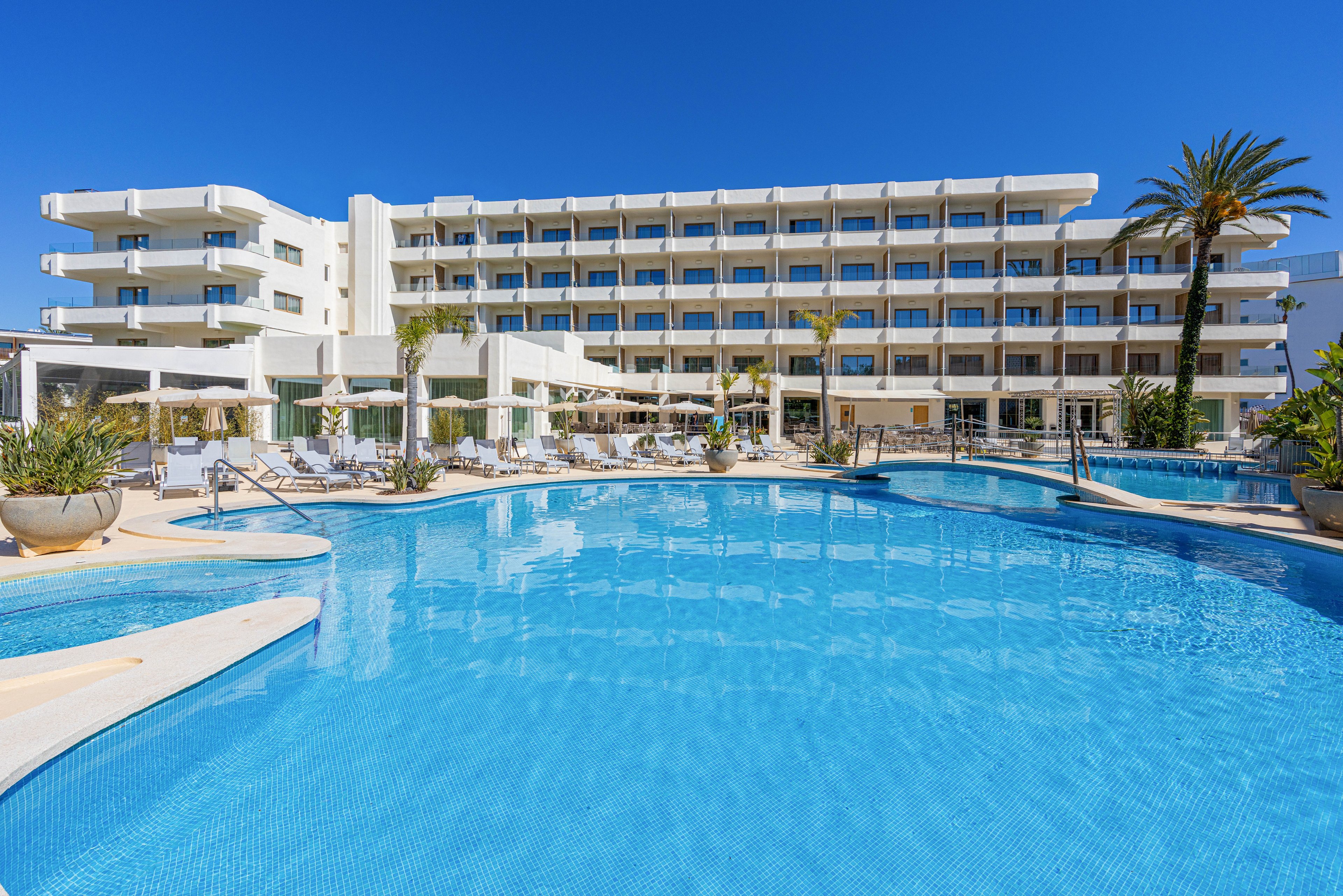 A contemporary multi-story resort hotel overlooks a large, curved outdoor swimming pool. Sun loungers and umbrellas line the poolside, suggesting a relaxing vacation setting. Palm trees and potted plants add a tropical touch to the bright, sunny scene. The clear blue sky and shimmering water emphasize a warm, inviting atmosphere ideal for leisure and holidays.